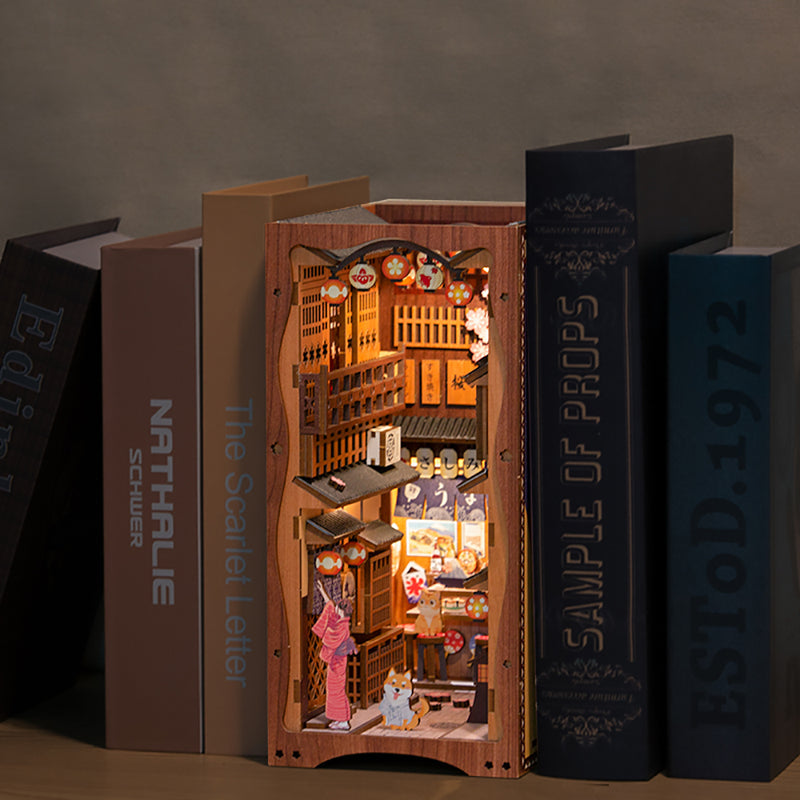 Under the Sakura Tree Book Nook (PREORDER) - Wooden miniature book nook lit with warm yellow lights, depicting a detailed Japanese street scene under blooming pink.