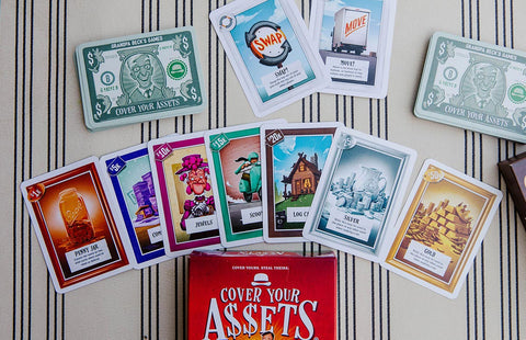Cover Your Assets - Colorful playing cards with various illustrations spread out above a red "Cover Your Assets" game box on striped fabric.