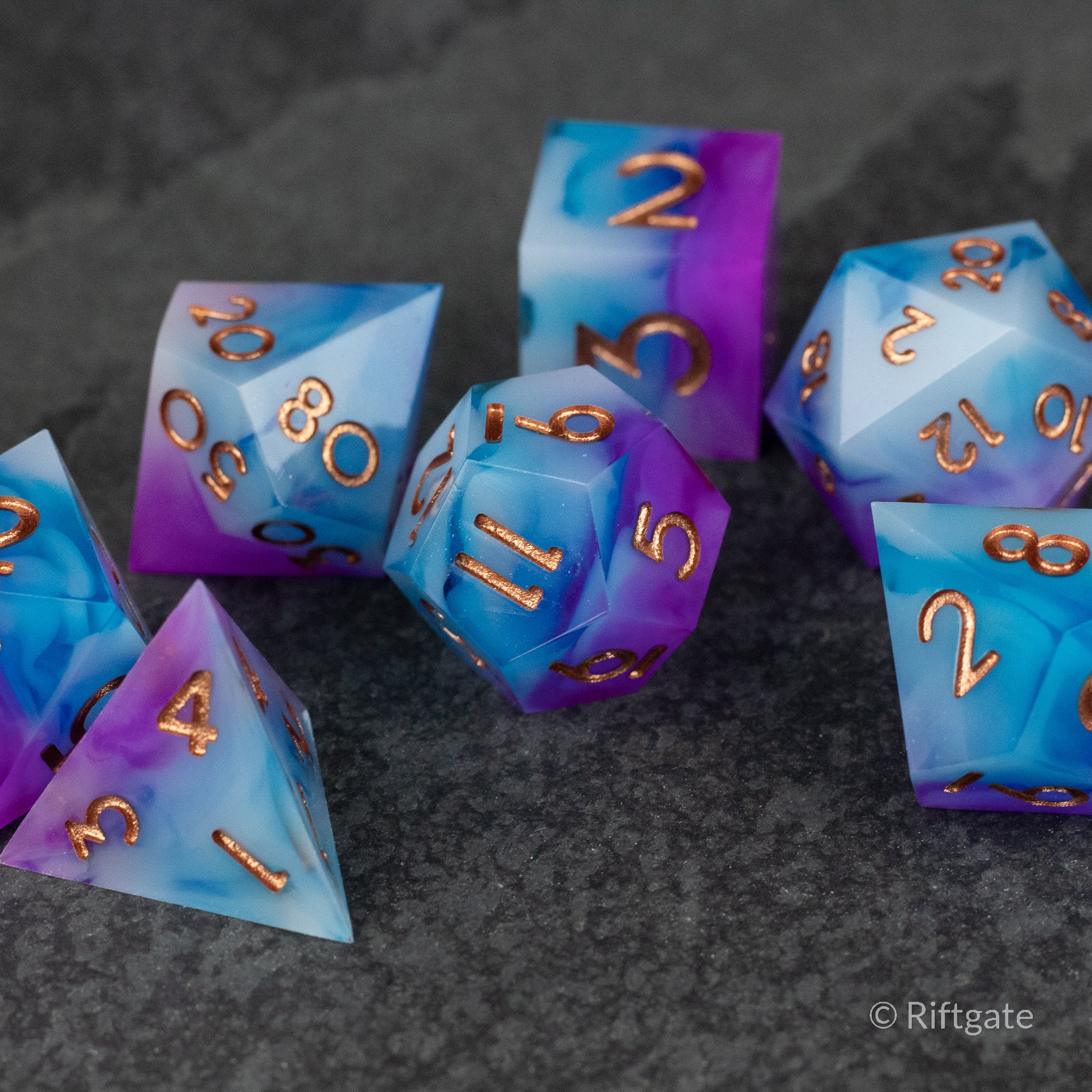 Arcane Tempest Dice Set - Set of seven sharp-edged polyhedral dice in a purple and blue gradient with metallic gold numbers on a dark textured surface.