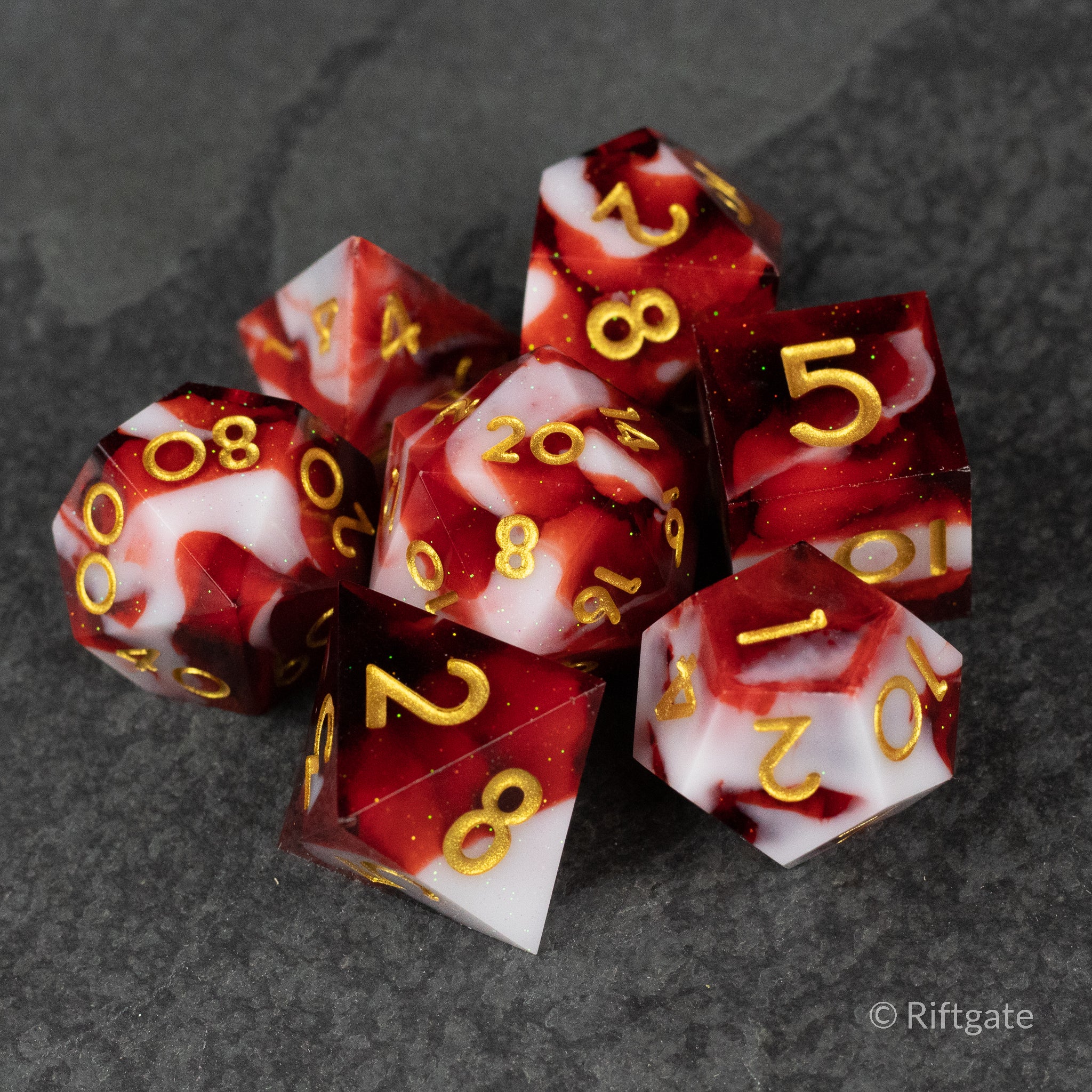 Blood and Snow Dice Set - Red and white marbled seven-piece sharp edge dice set with gold numbers on dark stone surface.