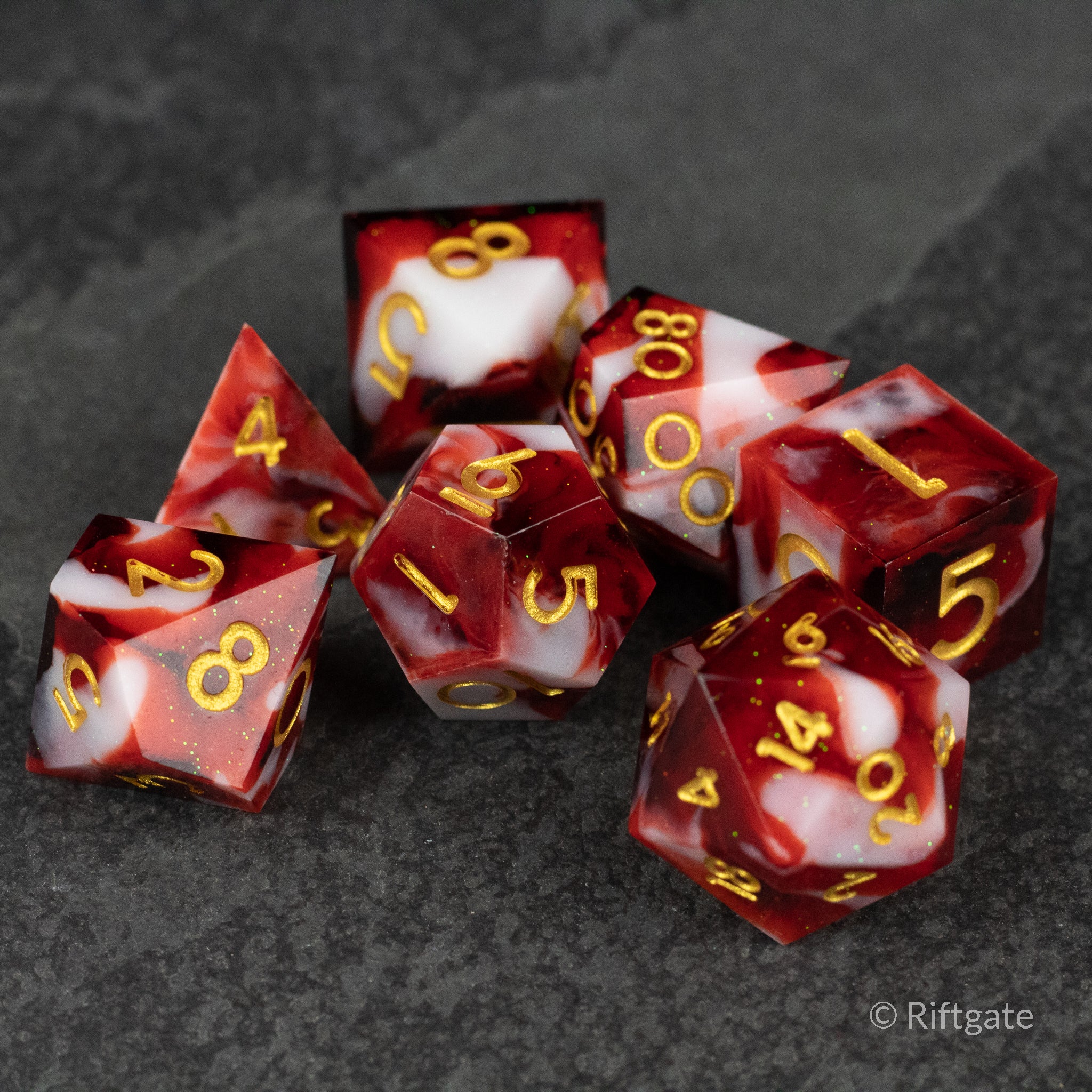Blood and Snow Dice Set - Seven sharp-edged polyhedral dice with swirling red and white marbled pattern and gold numbers on a dark gray surface.