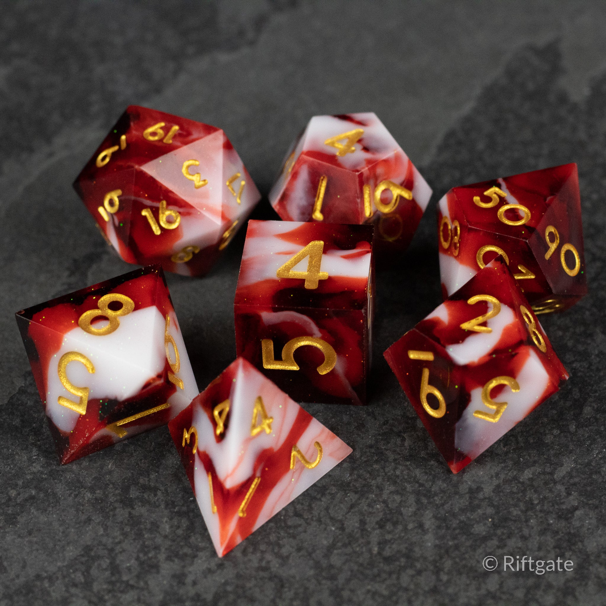 Blood and Snow Dice Set - Six glossy red and white marbled polyhedral dice with sharp edges and gold numbers on a dark textured surface.