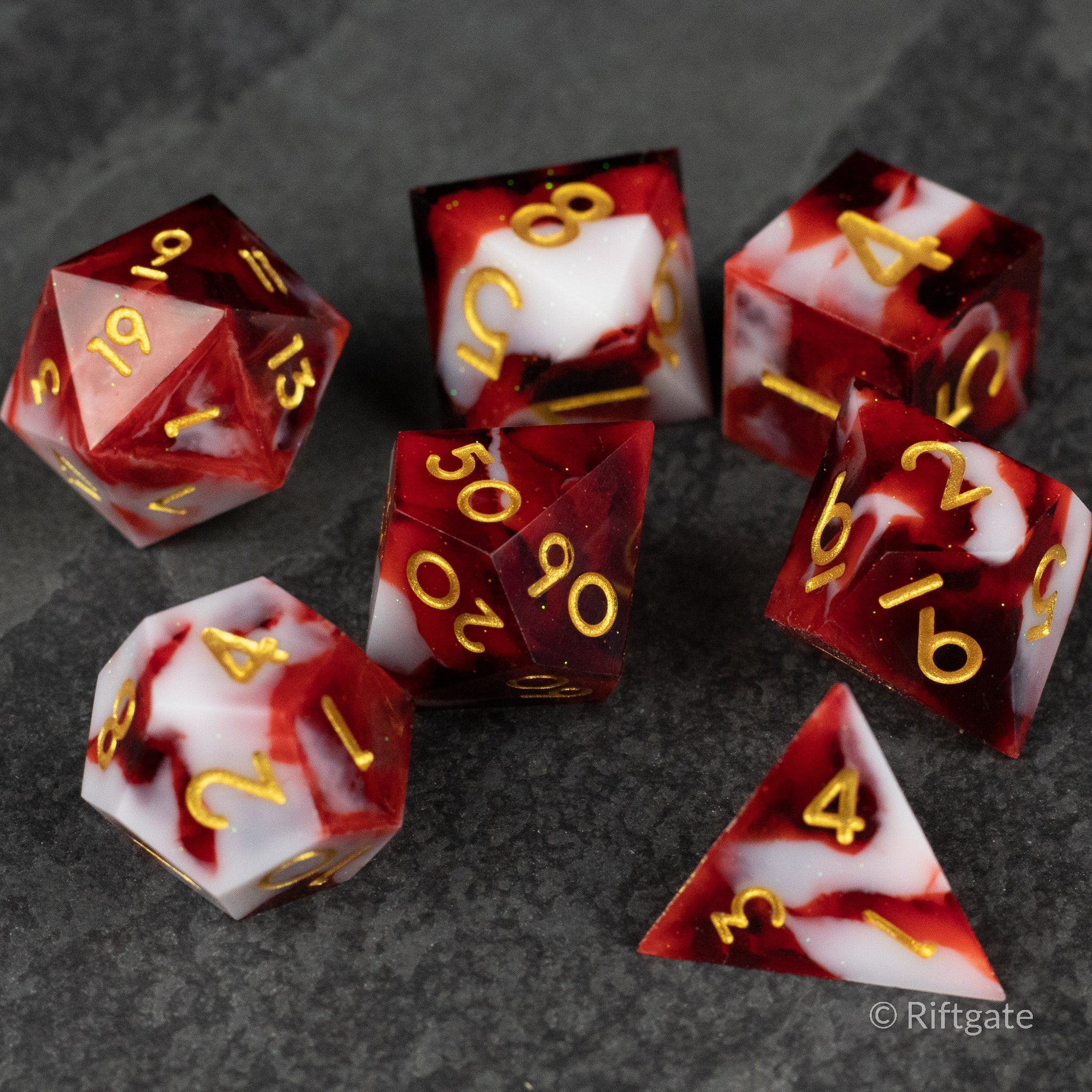Blood and Snow Dice Set - Seven sharp-edged polyhedral dice with red and white marbled resin and gold numbers on a dark background.