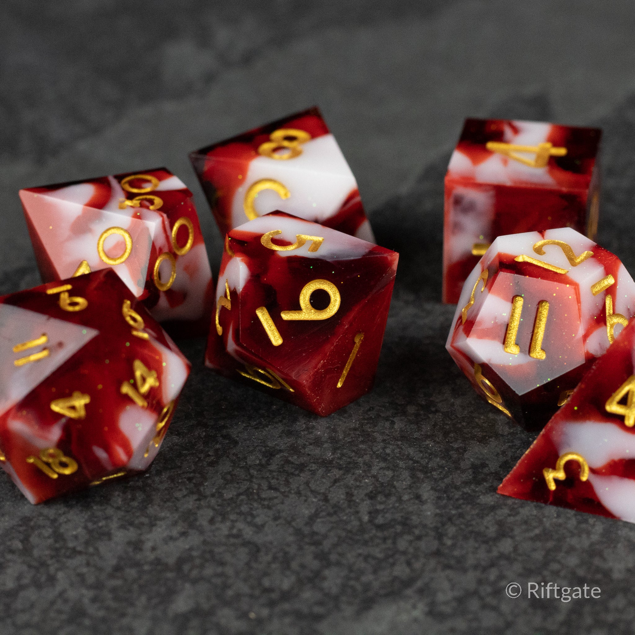 Blood and Snow Dice Set - Seven translucent red and white marbled polyhedral dice with sharp edges and gold numbers on a dark textured surface.