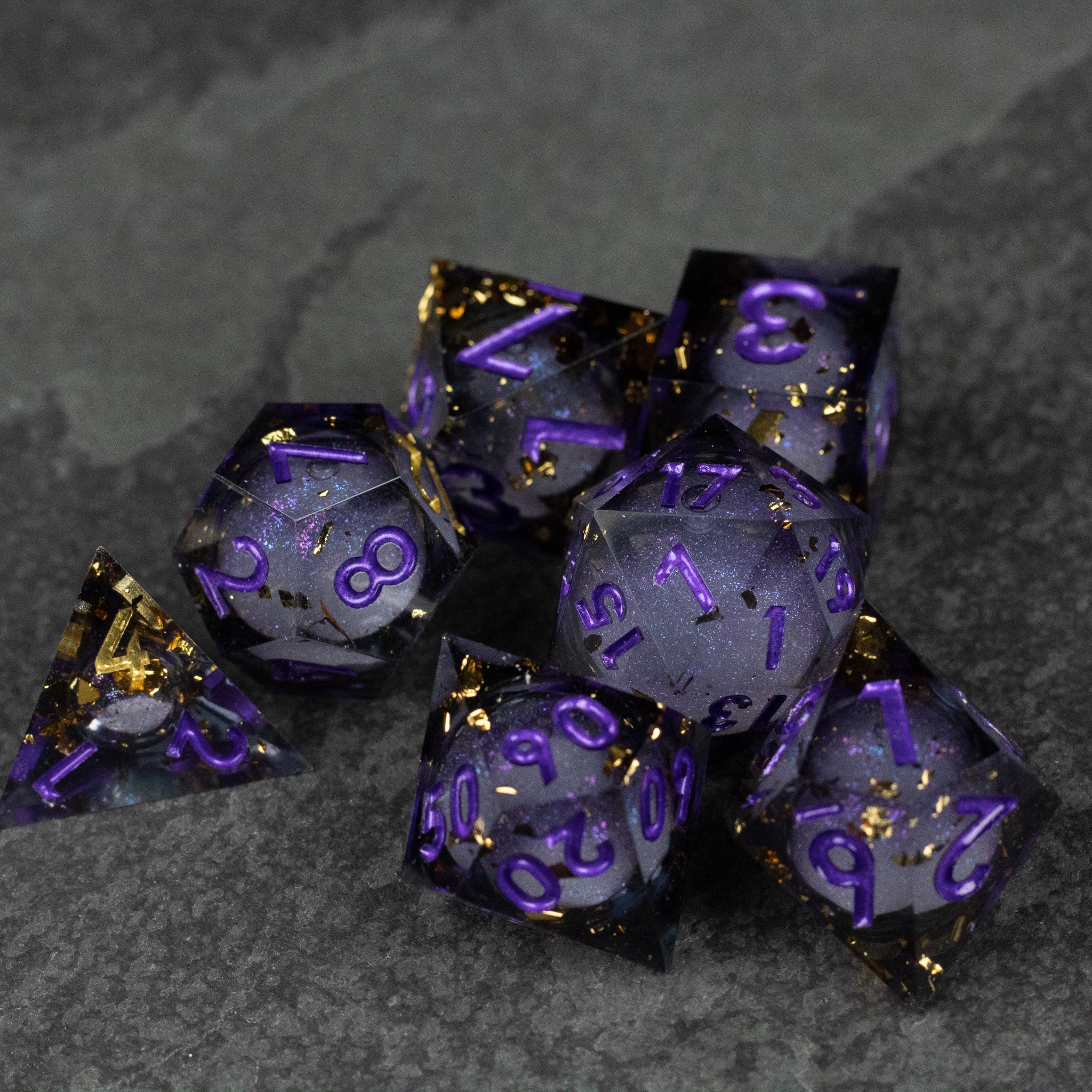 Witching Hour Liquid Core Dice - Set of seven translucent black sharp-edge dice with purple numbers and swirling gold liquid core on dark stone background.