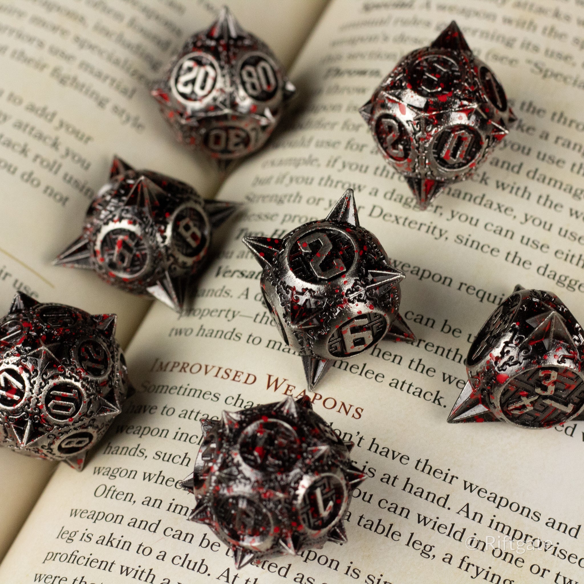 Bloody Morning Star Metal Dice Set - Seven black metal dice with red splatter and white numbering, shaped with sharp edges and spikes, arranged on an open book.