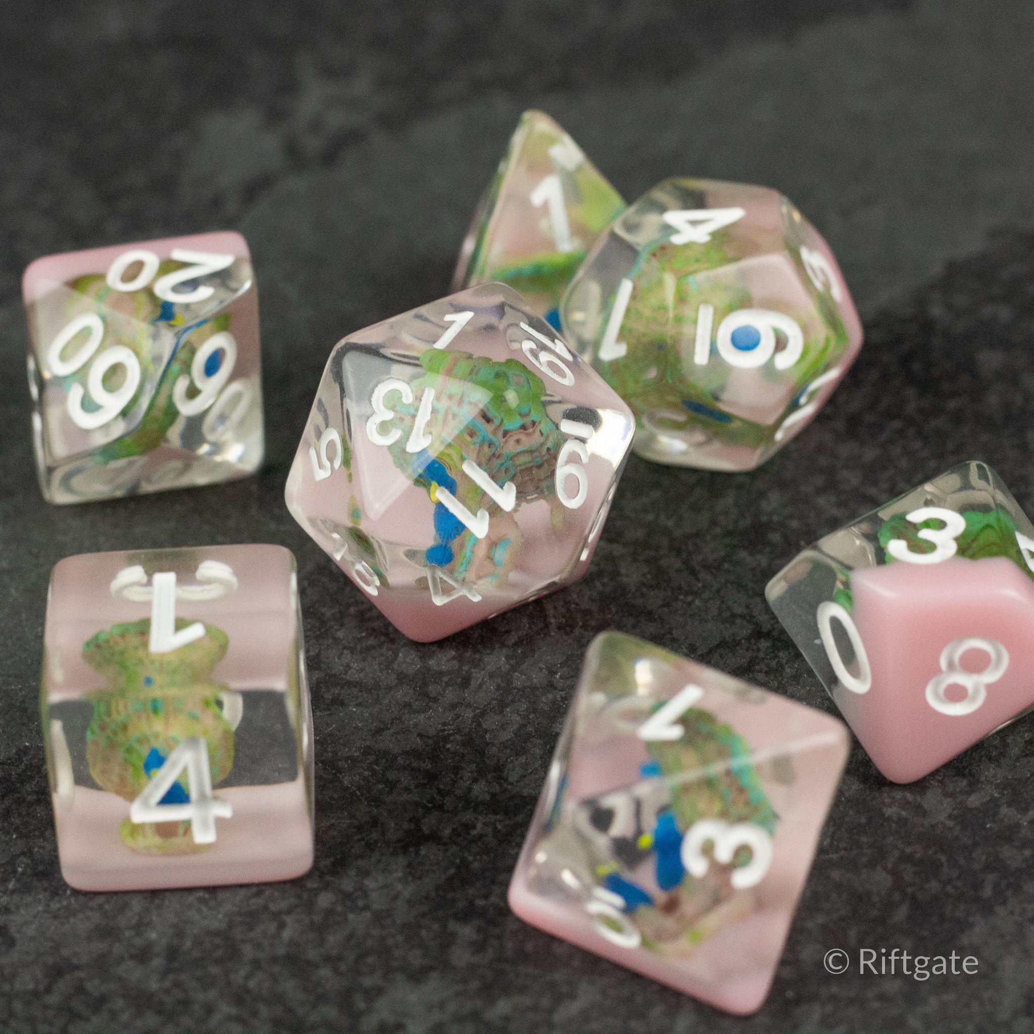 Peacock Dice Set - Seven transparent pink polyhedral dice with green, blue, and yellow flecks and white numbers on a dark surface.