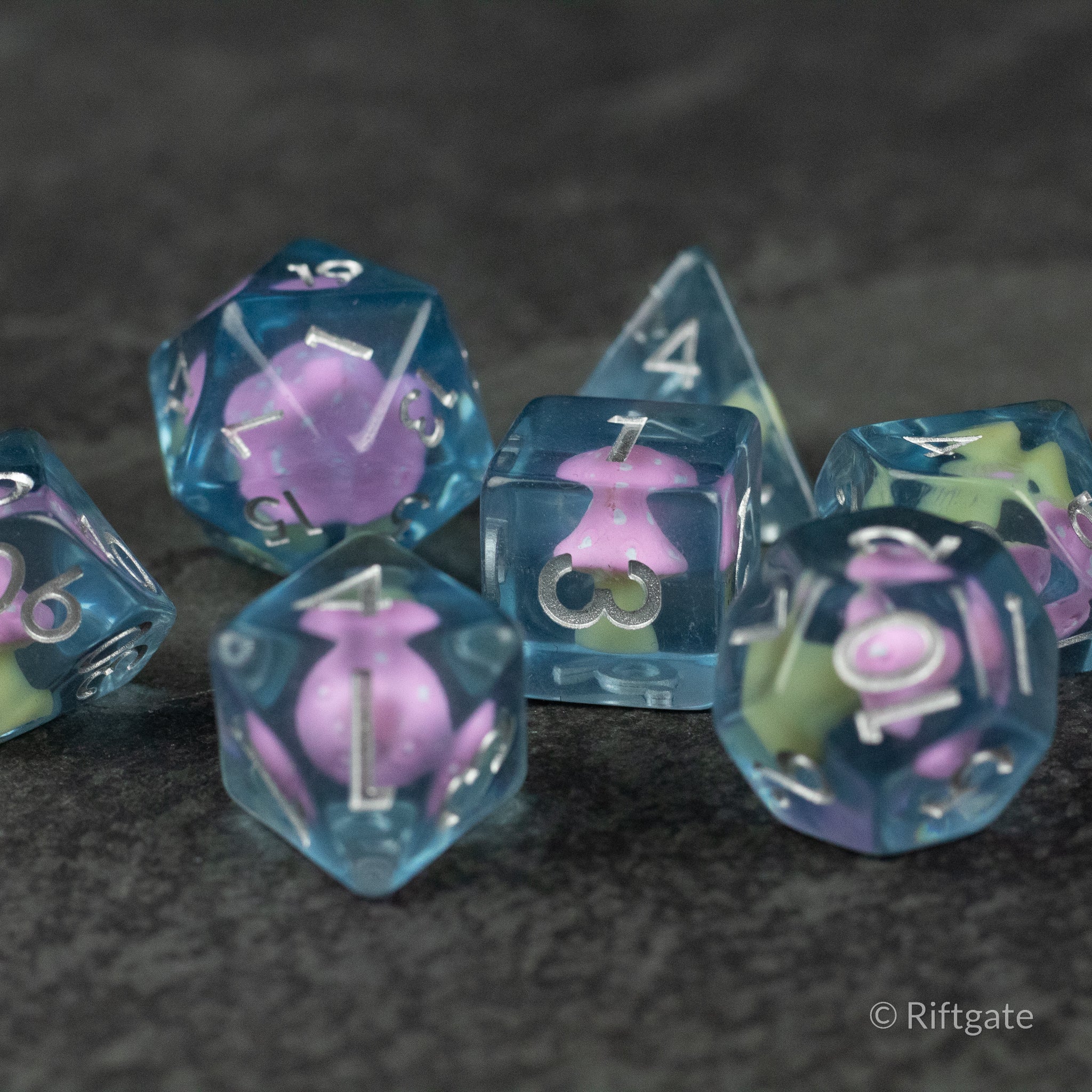 Pink + Blue Resin Mushroom Dice Set - Blue translucent resin dice set with embedded pink and light green mushroom inclusions on dark background.