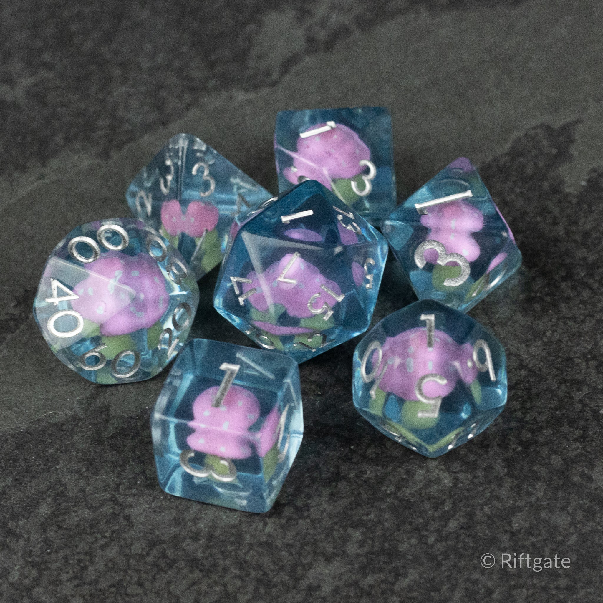 Pink + Blue Resin Mushroom Dice Set - Seven blue resin dice with pink mushroom inclusions and white numbers, including d20, d12, d10, d8, d6, and d4 shapes.