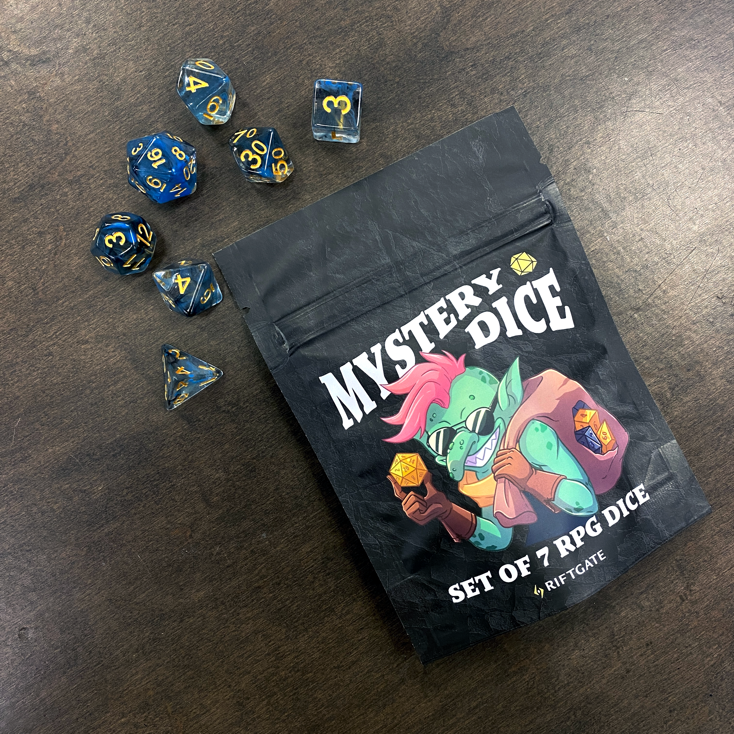 Dice Booster Pack - Set of seven dark blue and gold RPG dice spilling from a black pouch labeled "Mystery Dice" with colorful fantasy artwork on