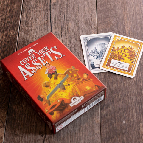 Cover Your Assets - Cover Your Assets card game box with red cover and two visible cards featuring gold and silver assets on wooden floor.