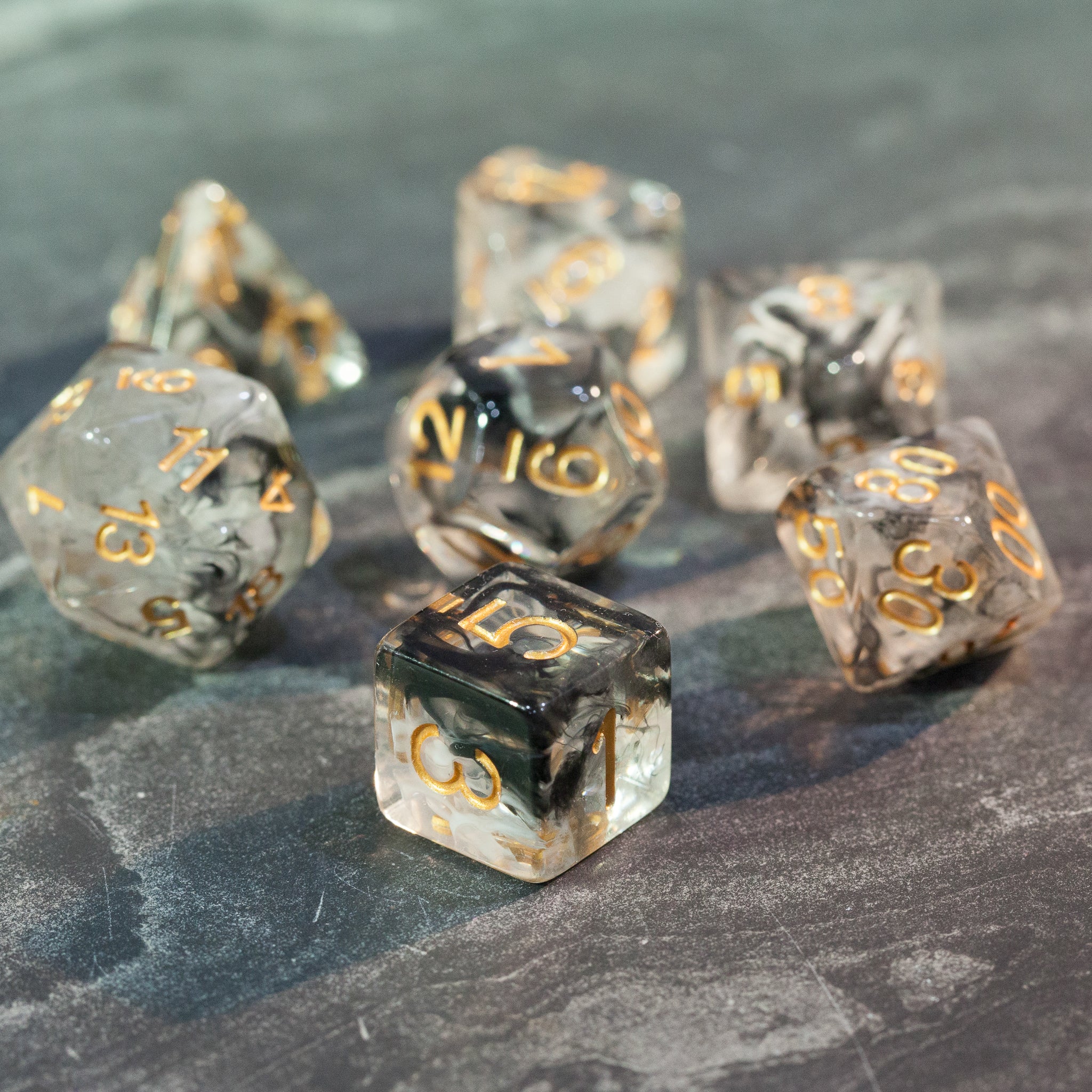 Black & White Smoke Dice Set - Seven black and white smoke-effect polyhedral dice with gold numbers on a gray surface.