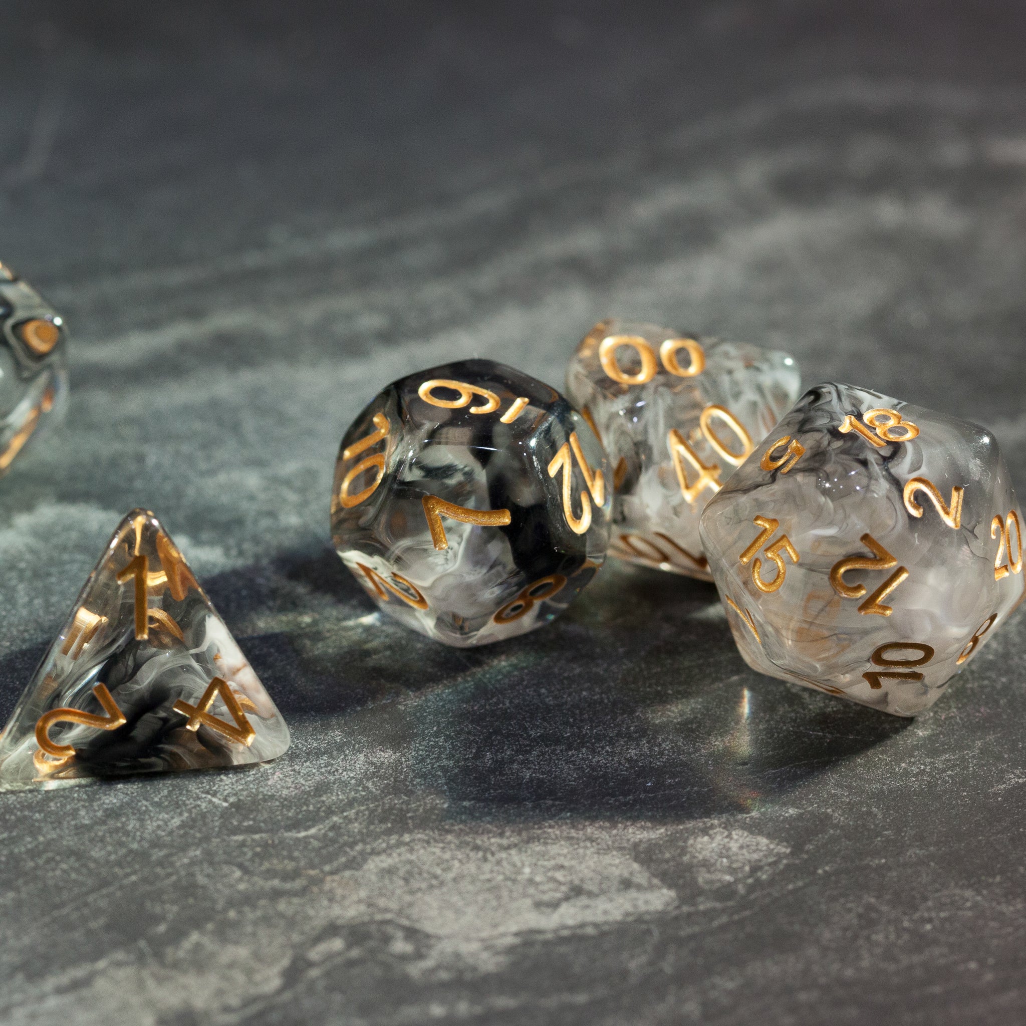 Black & White Smoke Dice Set - Set of translucent black and white smoky polyhedral dice with gold numbering on gray surface.