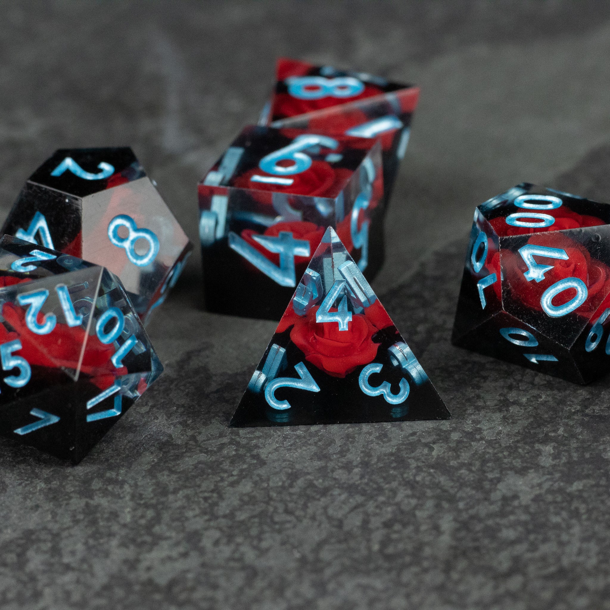 Cursed Rose Dice Set - Black and red sharp-edged dice set with blue numbers, featuring a d20, d12, d10, d8, d6, and d4 on a gray surface.