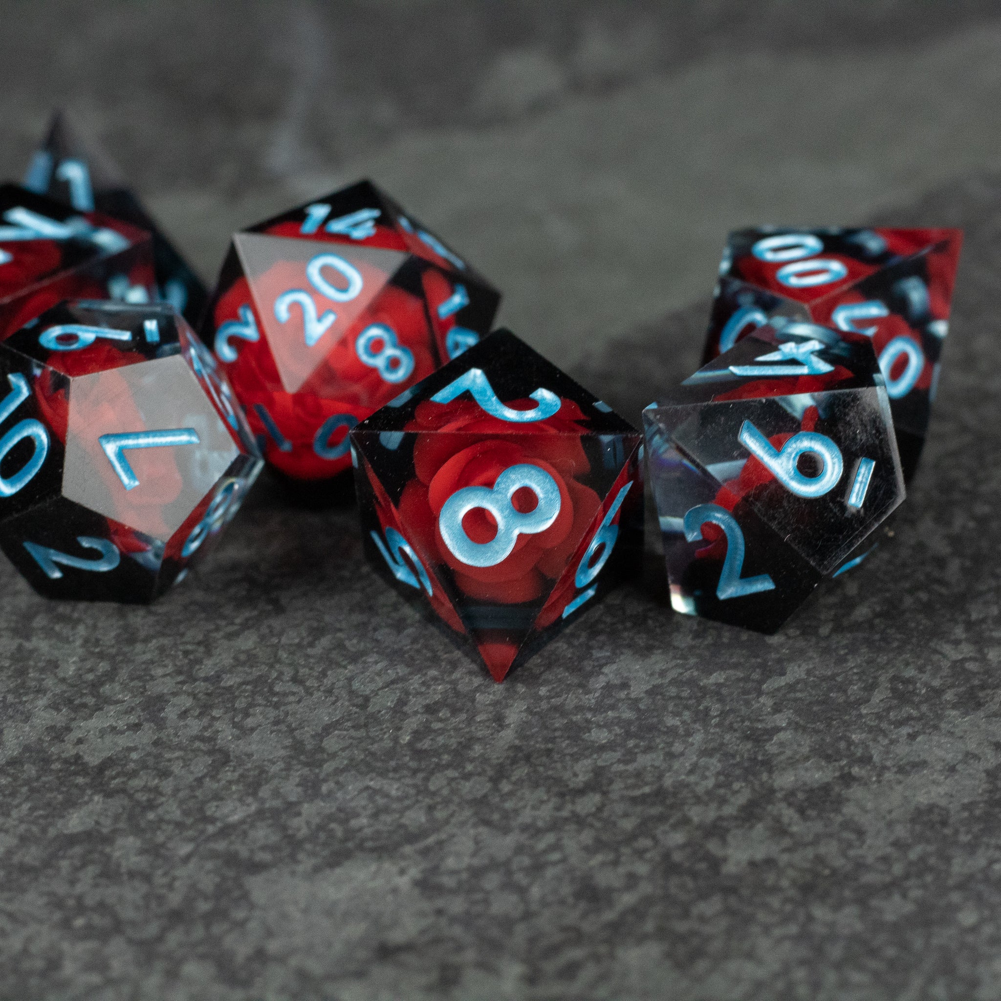 Cursed Rose Dice Set - Red and black marbled sharp edge polyhedral dice with light blue numbers on gray surface.