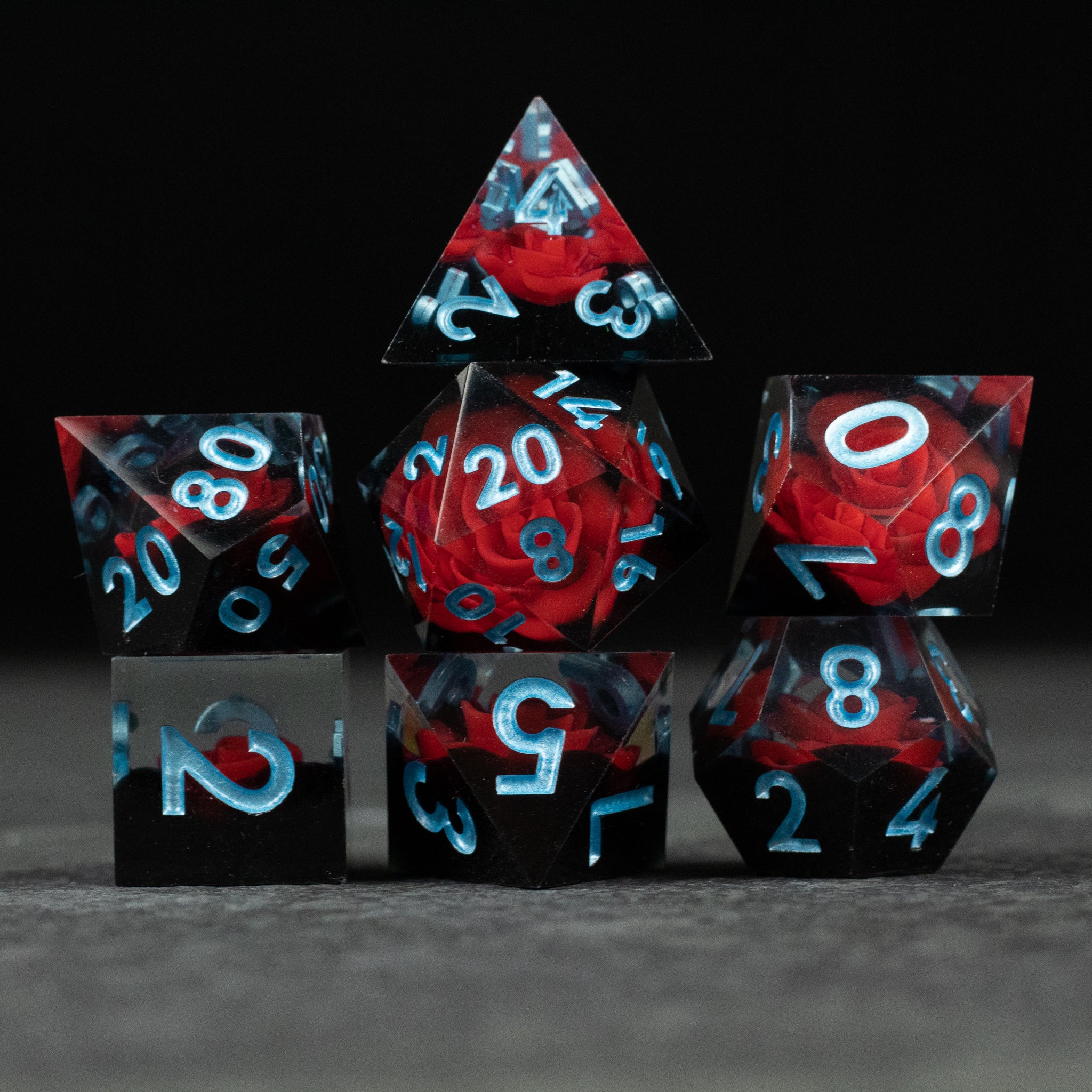 Cursed Rose Dice Set - Seven polyhedral dice with sharp edges, translucent red and black marbled with blue numbers, featuring a visible red rose.