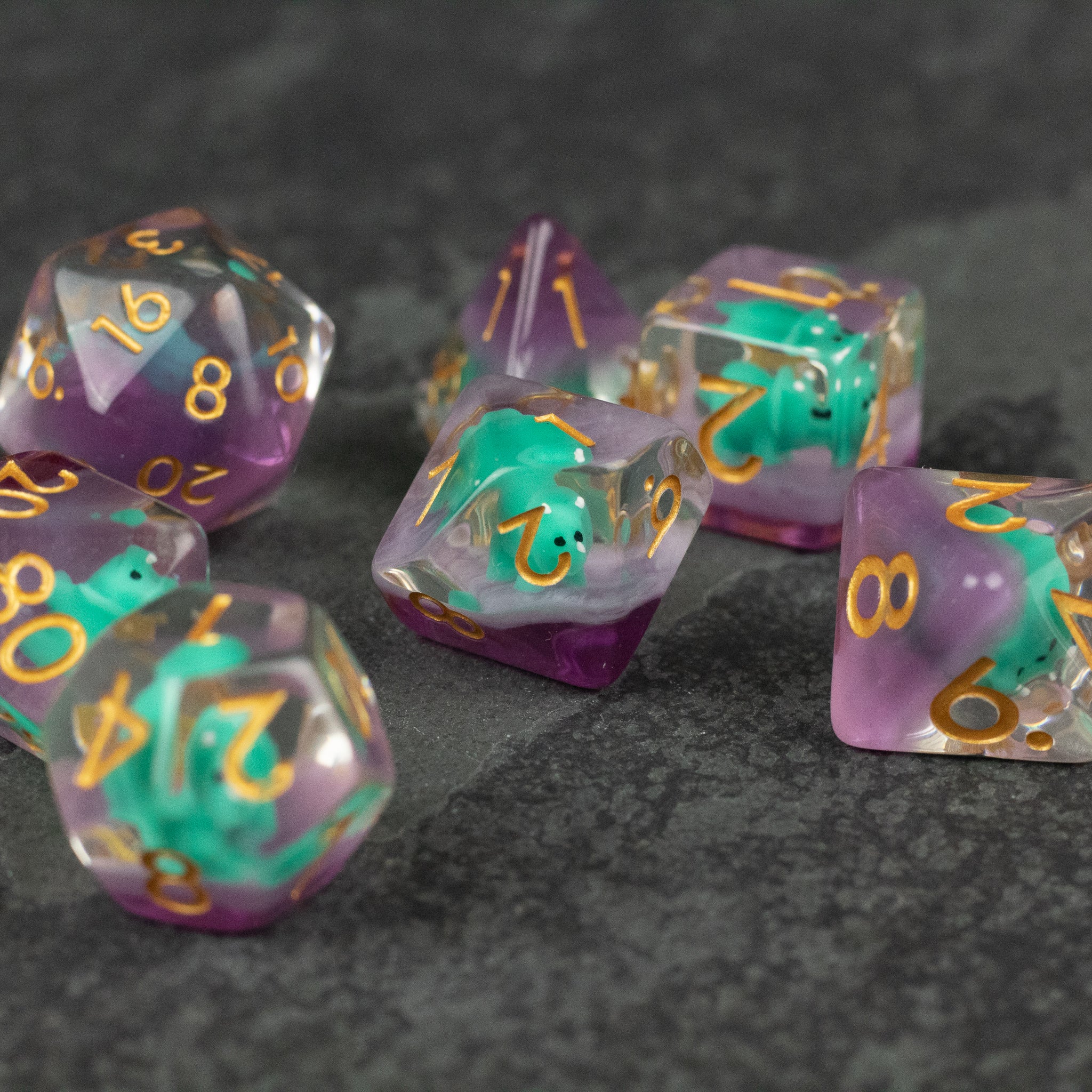 Green Triceratops Dice Set - Translucent purple and green polyhedral dice with gold numbering, including d20 and d6 shapes on a dark textured surface.