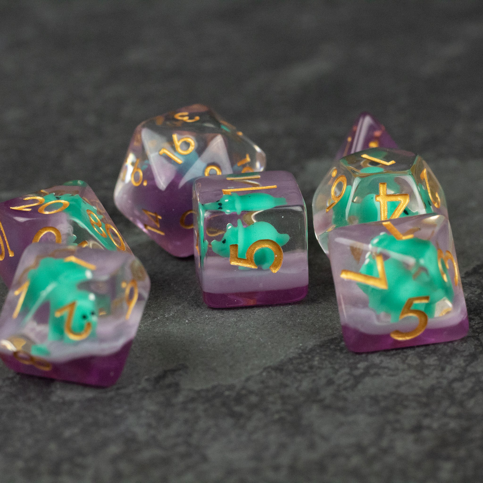 Green Triceratops Dice Set - Set of translucent purple and green polyhedral dice with gold numbers on dark textured surface.