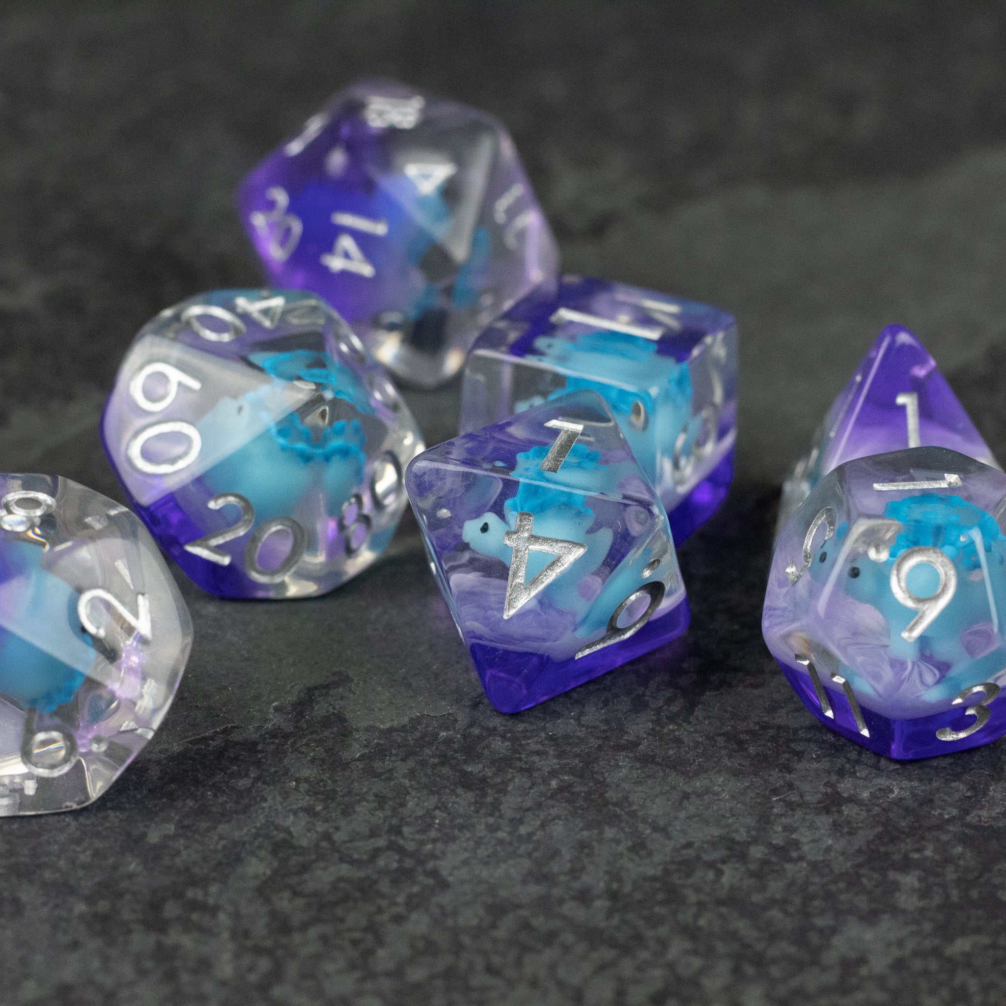 Blue Stegosaurus Dice Set - Set of seven translucent polyhedral dice in varying shades of blue and purple with white numbering on a dark textured.