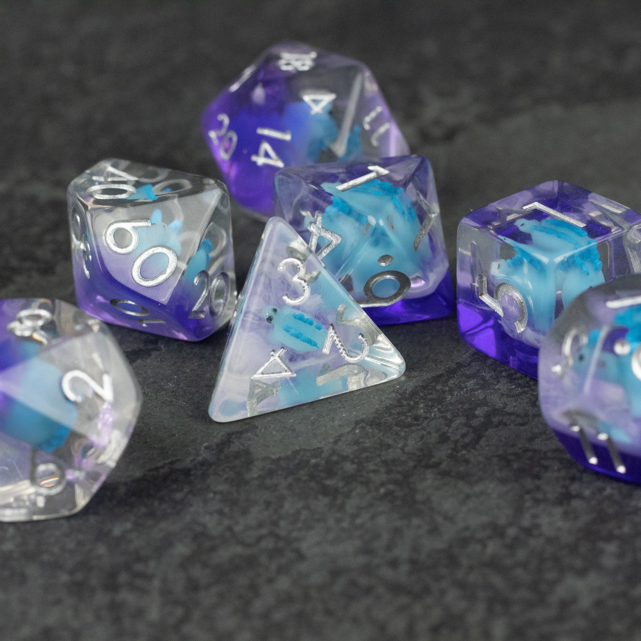 Blue Stegosaurus Dice Set - Blue and purple translucent polyhedral dice set with white numbering, featuring a d4, d6, d8, d10, d12, and d20 on a dark.