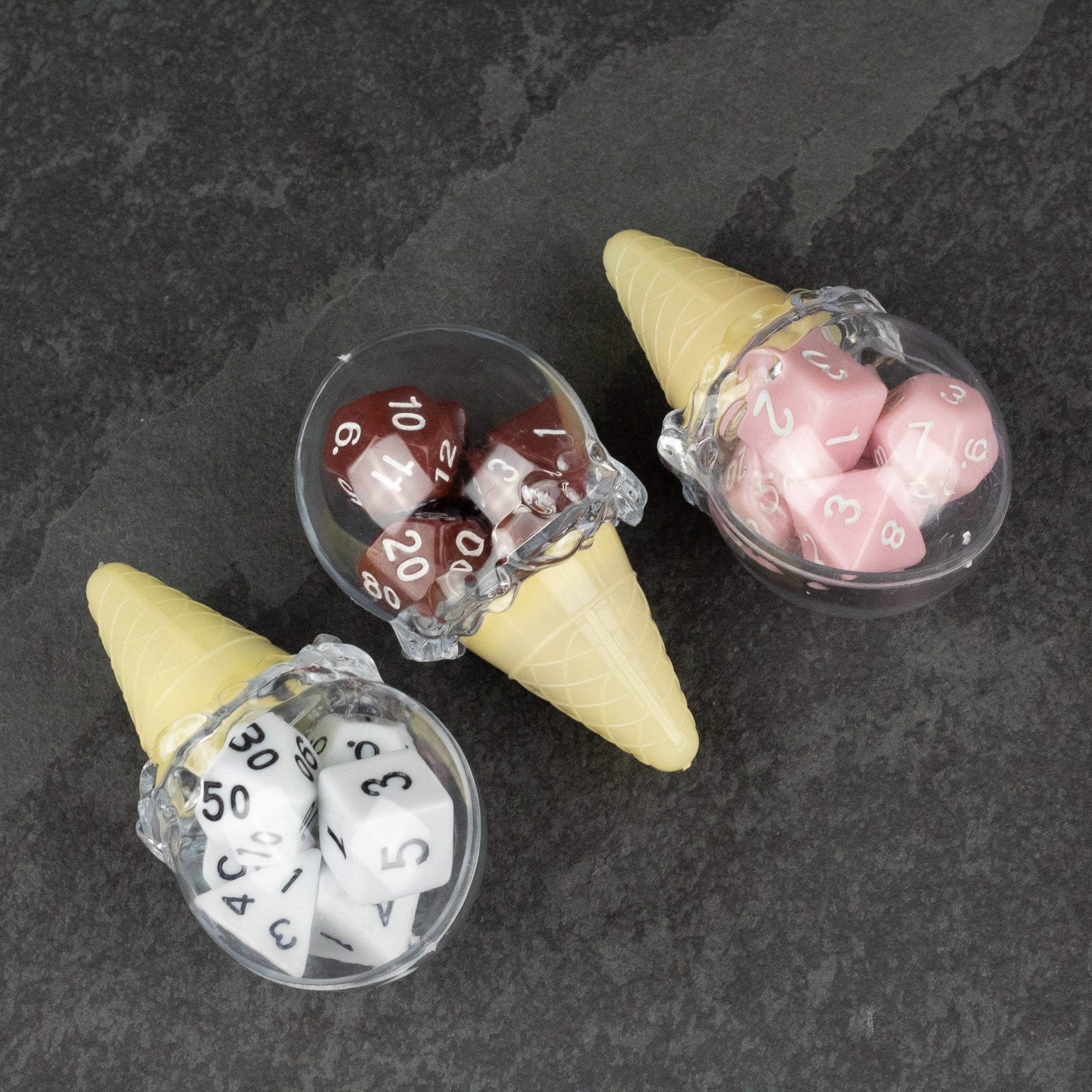 Dice Cream Cones - Three ice cream cone-shaped containers holding polyhedral dice sets in white, burgundy, and pink colors on a dark surface.