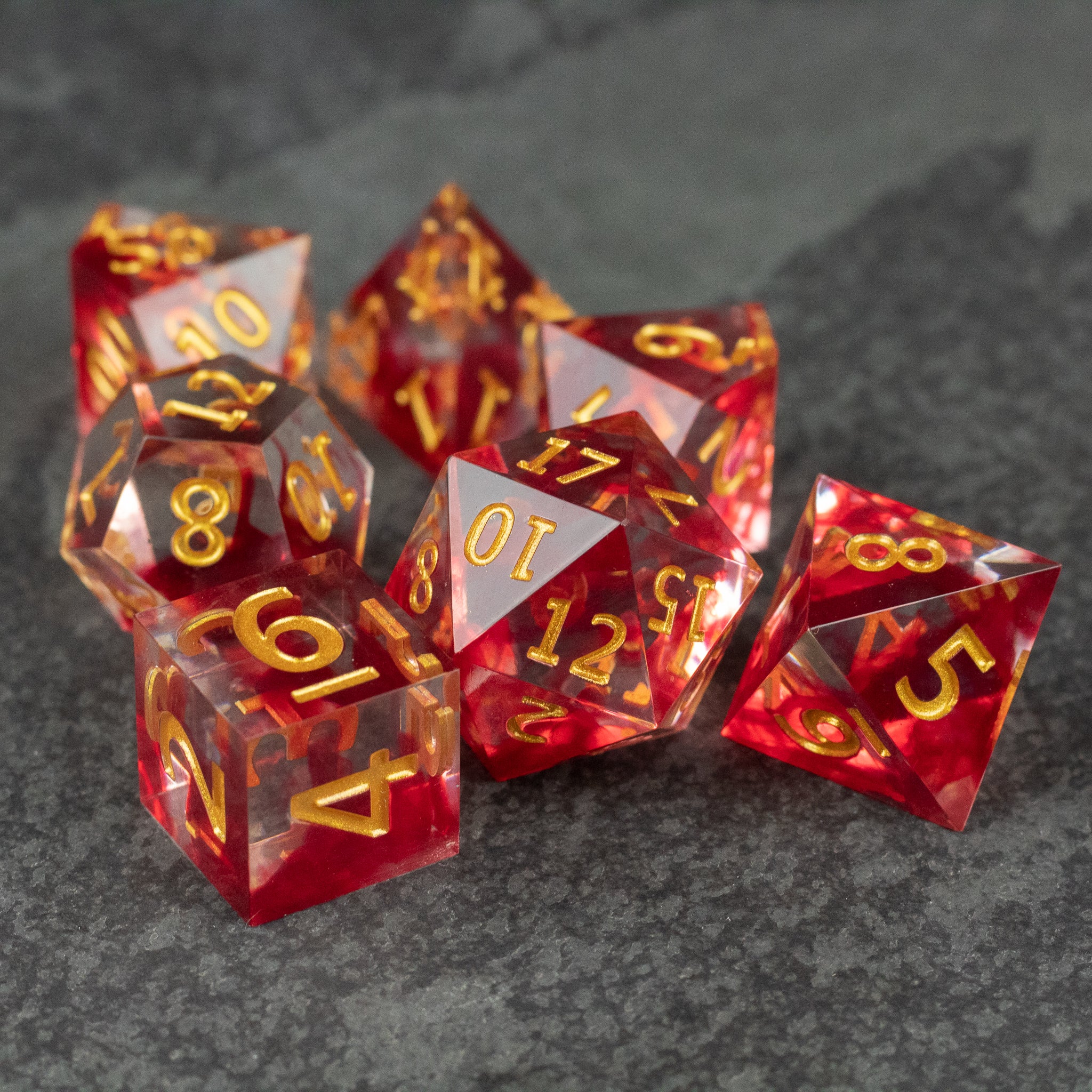 Blood Pact Dice Set - Red translucent sharp-edged polyhedral dice set with gold numbers on dark stone surface.