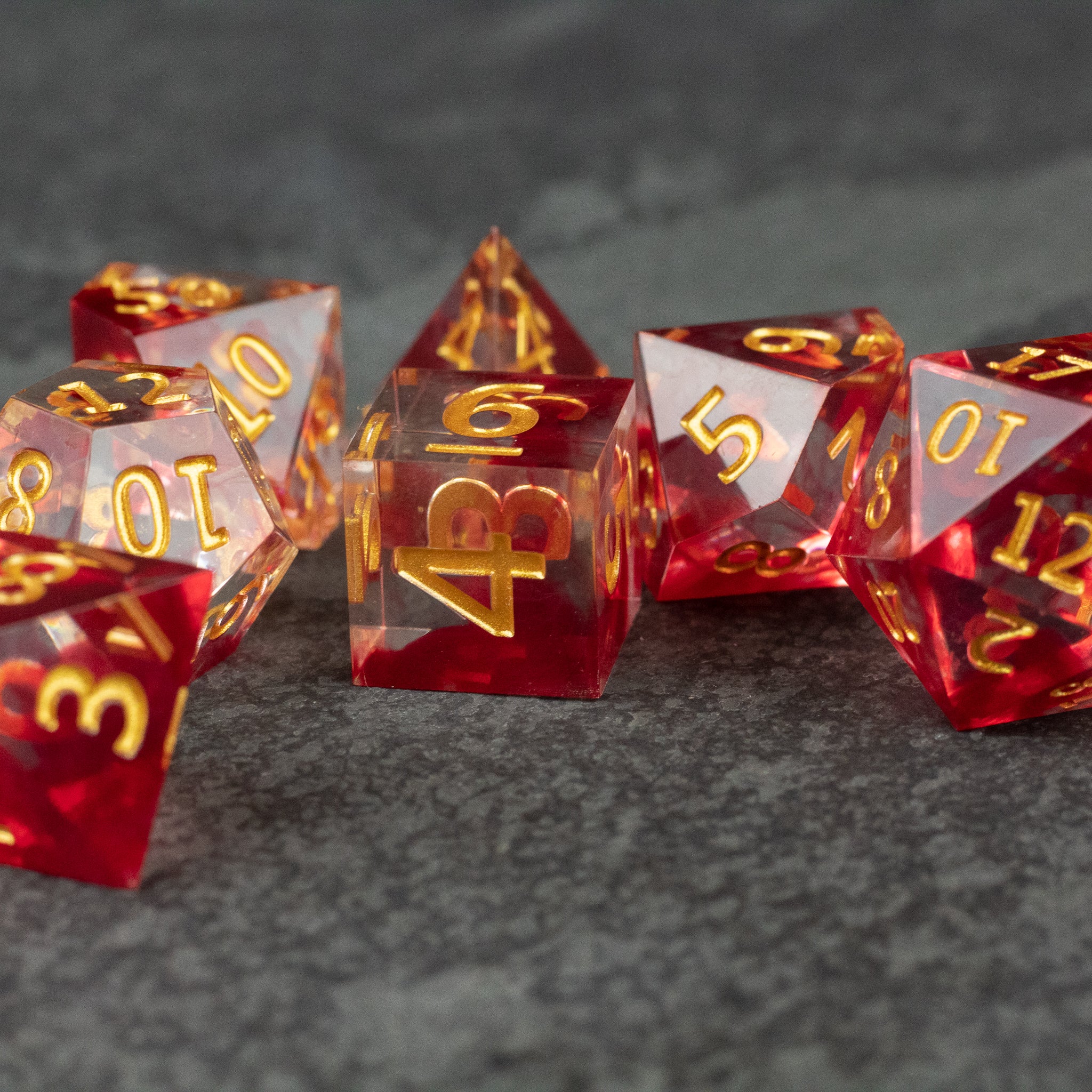Blood Pact Dice Set - Translucent red sharp-edged polyhedral dice set with gold numbering on a gray surface.