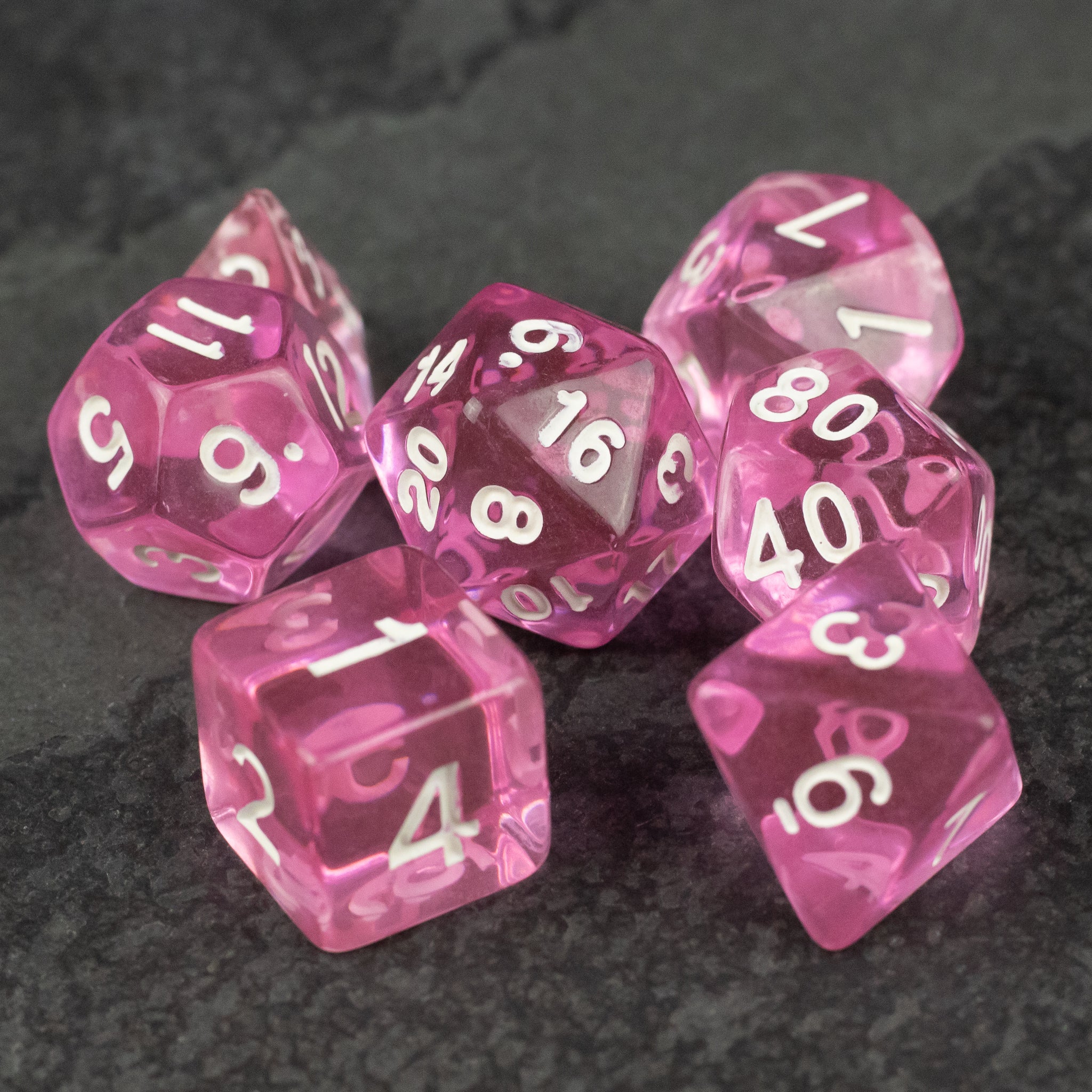 Pink Transparent Dice Set - Set of seven pink transparent polyhedral dice with white numbers on dark surface.