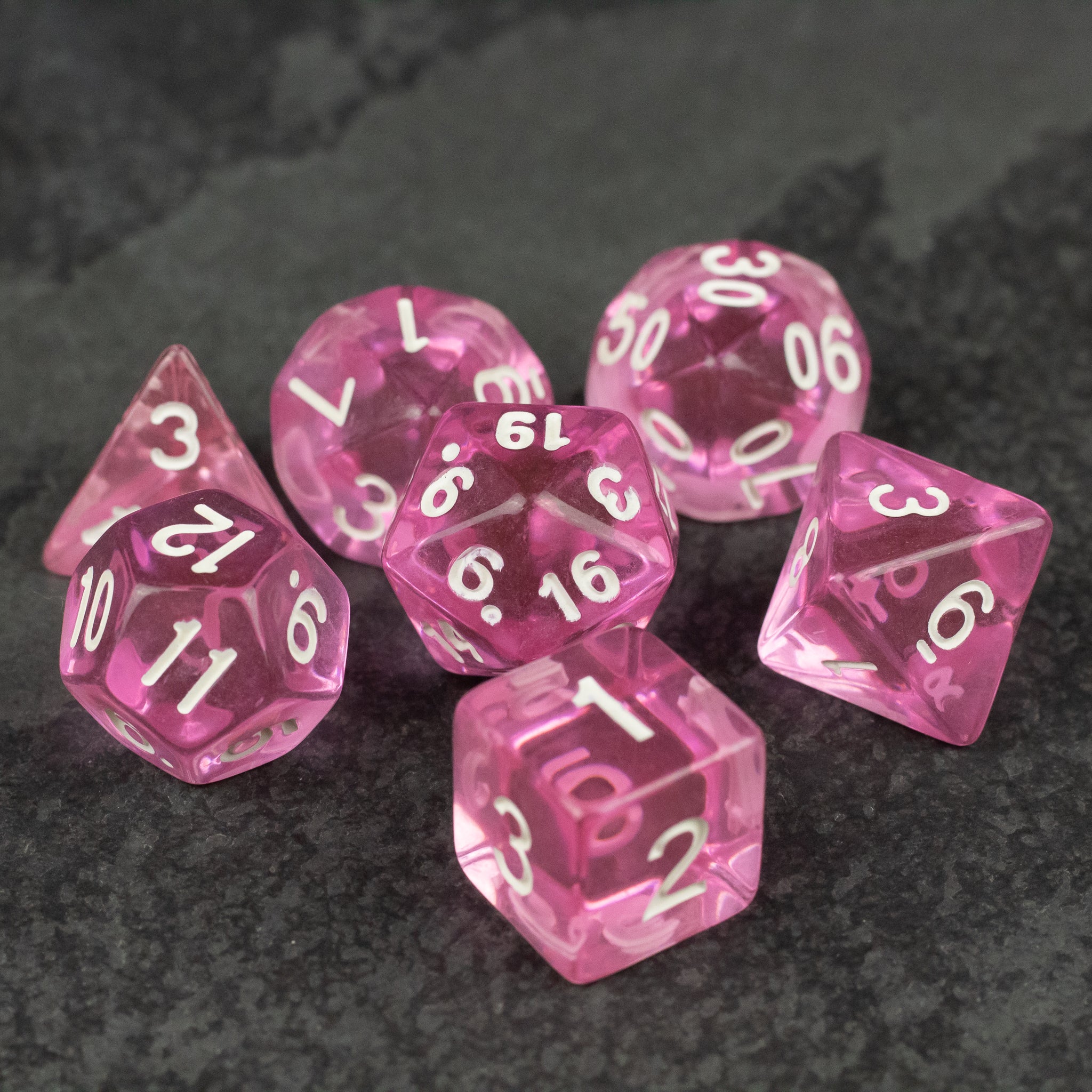 Pink Transparent Dice Set - Set of seven pink transparent polyhedral dice with white numbering on dark textured surface.