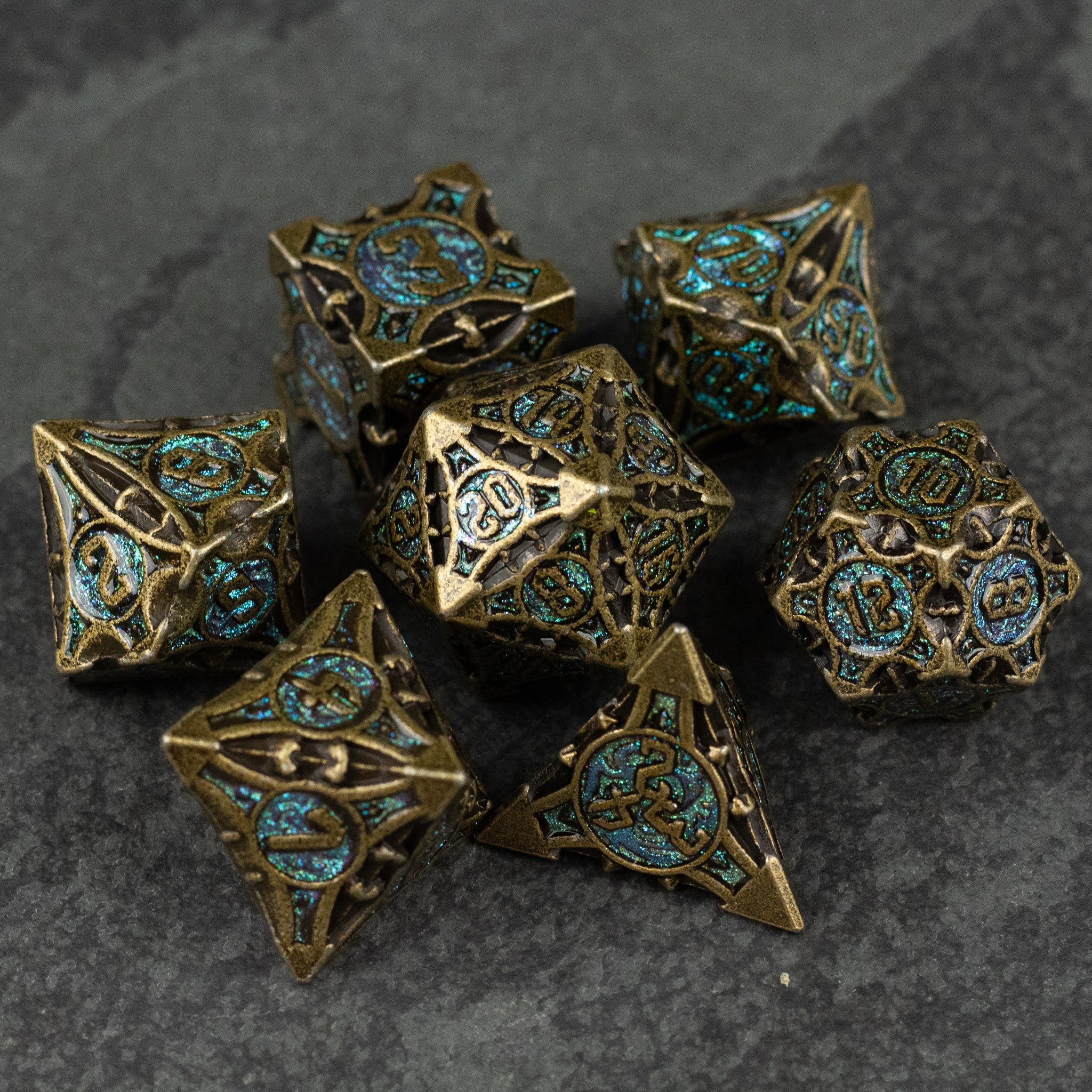 Empyrean Artifact Dice Set - Seven ornate metal polyhedral dice with intricate filigree designs and turquoise inlays on a dark stone surface.
