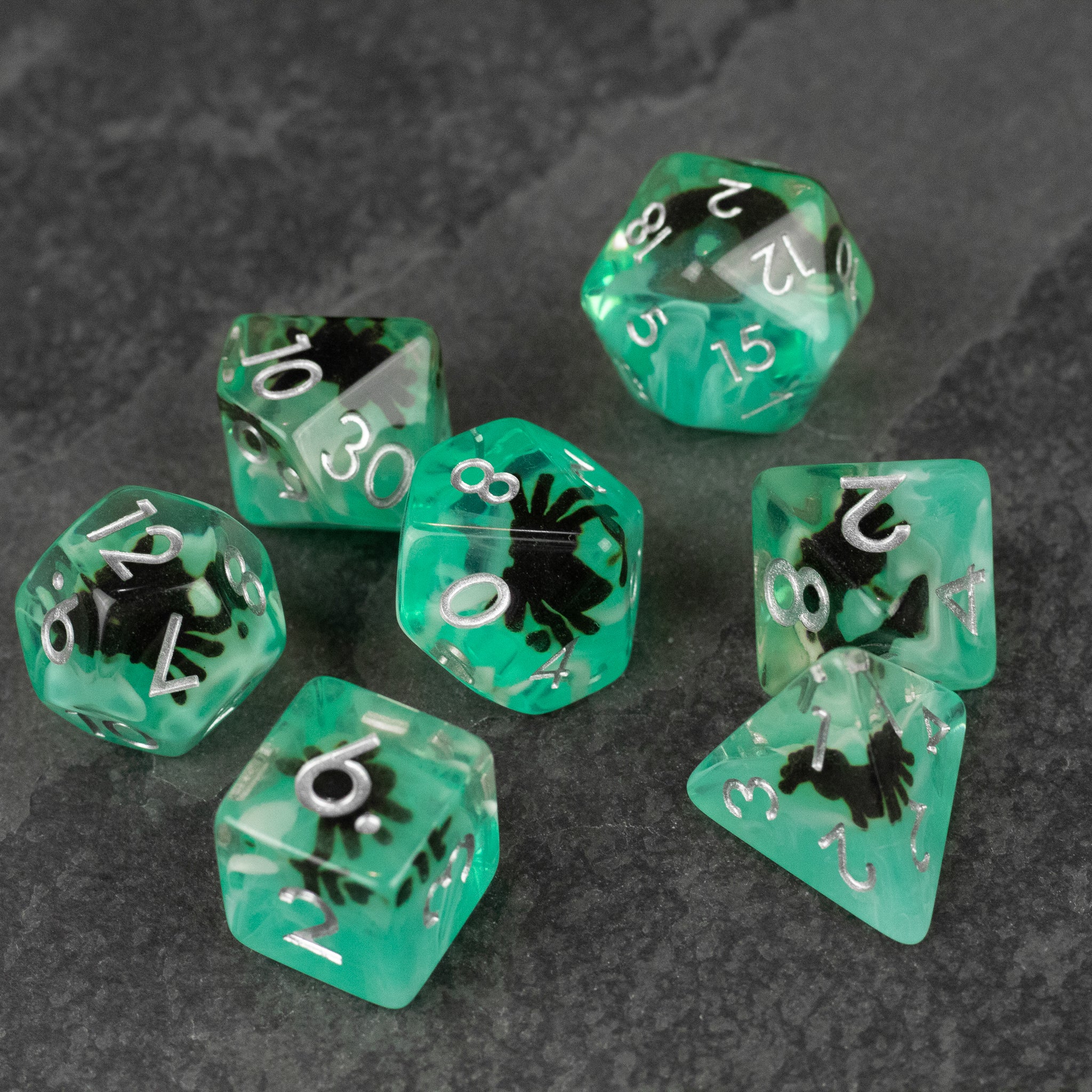 Black Spider Dice Set - Green - Seven green translucent polyhedral dice with black spider silhouettes and white numbers on a dark stone background.