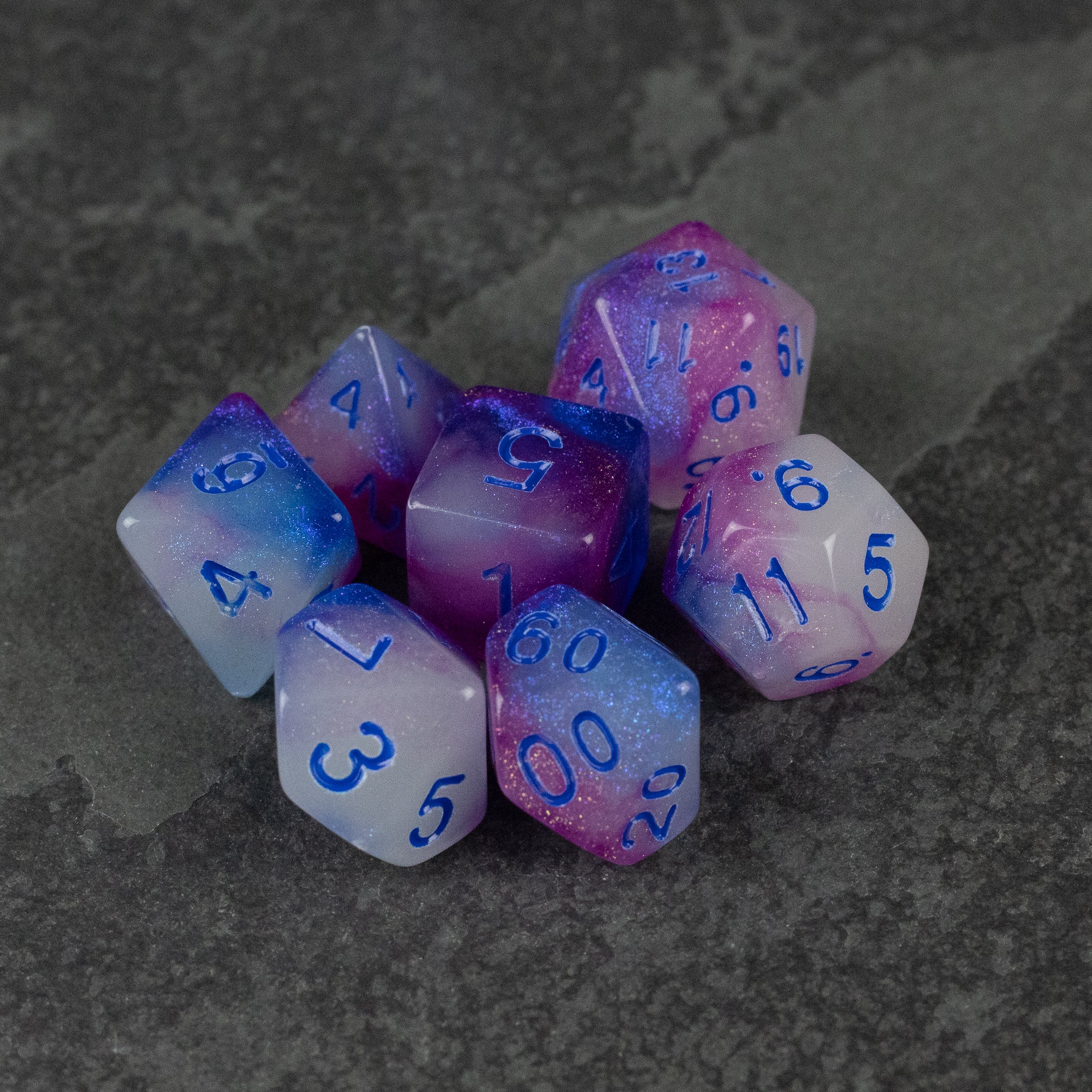 Emberlight Twilight Glow-in-the-Dark Dice Set - Set of seven purple, pink, and white gradient glow-in-the-dark RPG dice with blue numbers on a dark textured surface.