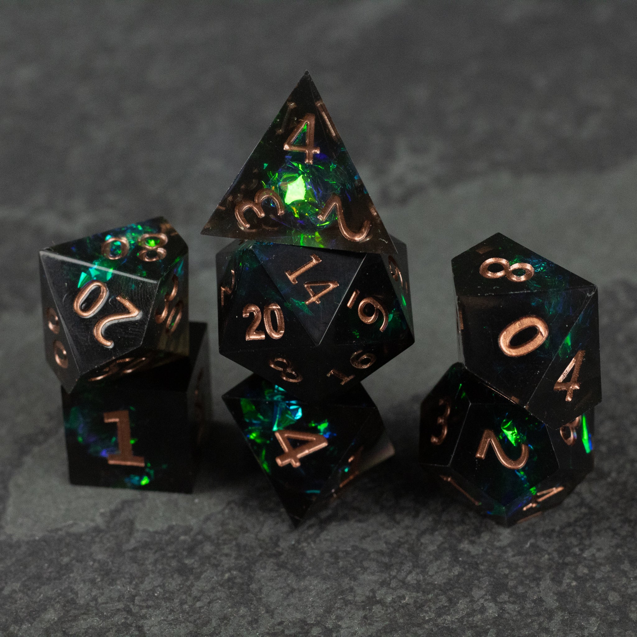 The Underdark Dice Set - Black translucent polyhedral dice set with sharp edges, glowing green and blue inclusions, and copper-colored numbering.