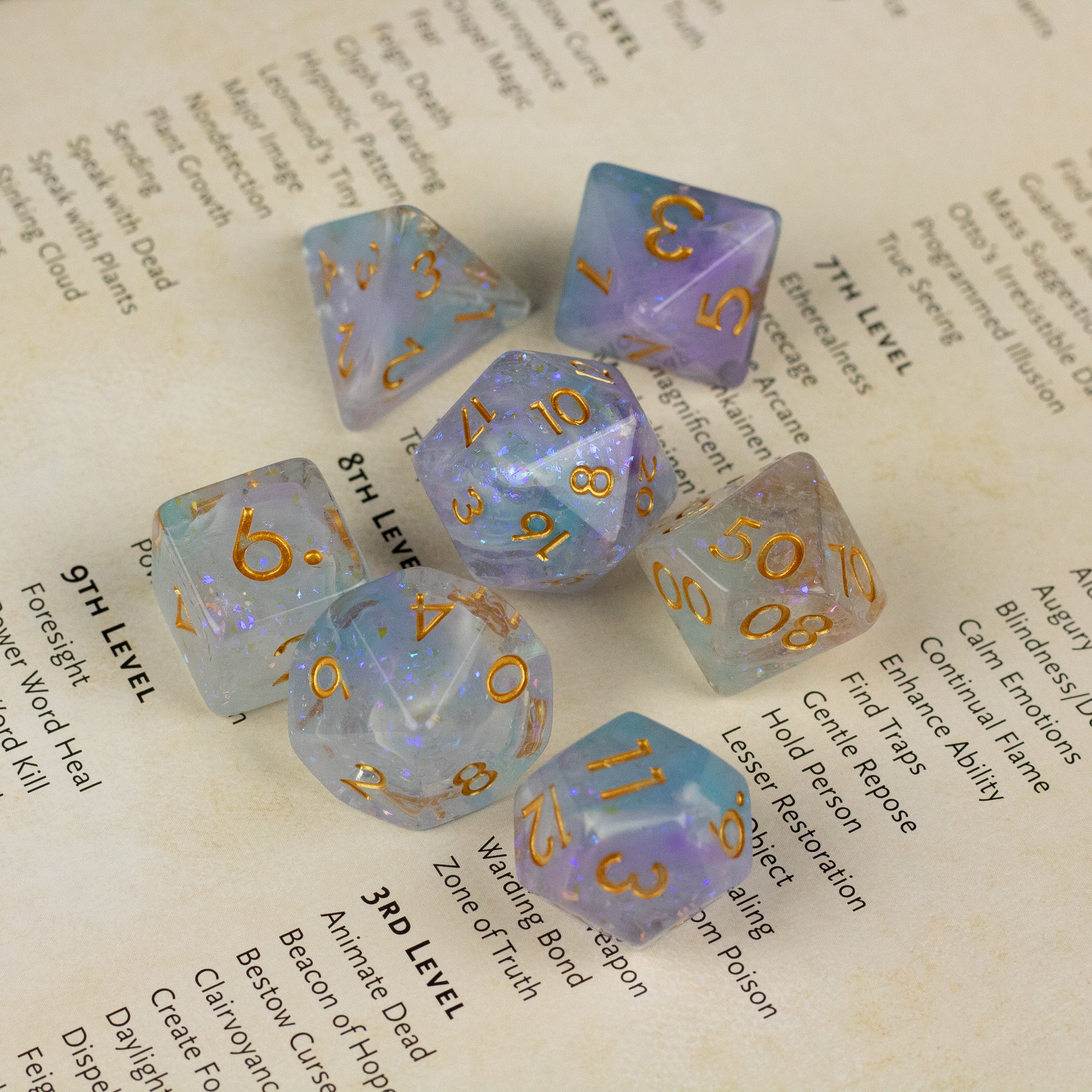 Spellbound Dice Set - Seven translucent dice with a gradient of purple and gold flakes, featuring gold numbers in various polyhedral shapes.