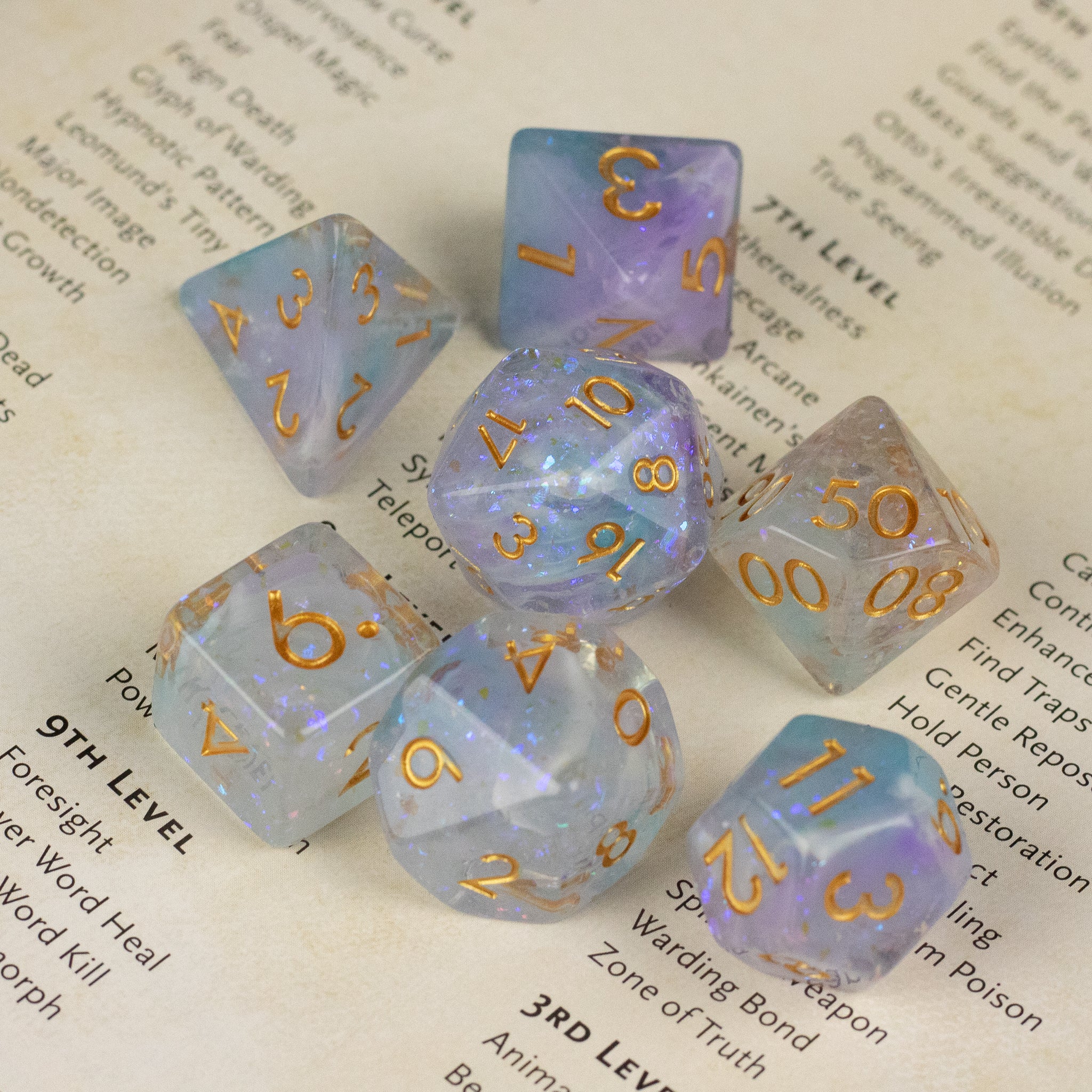 Spellbound Dice Set - Seven translucent dice with swirling blue and white colors and gold numbering on an open book page.