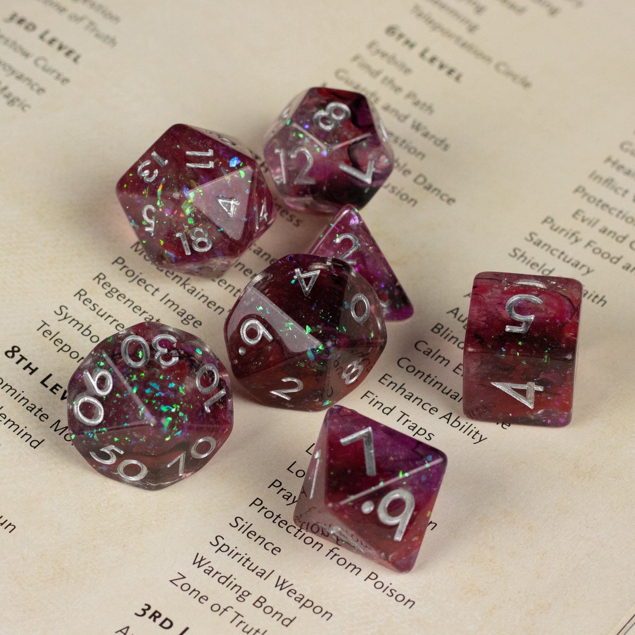 Black and red Spellweaver Dice Set - Seven black and red translucent polyhedral dice with white numbers scattered on a beige background.