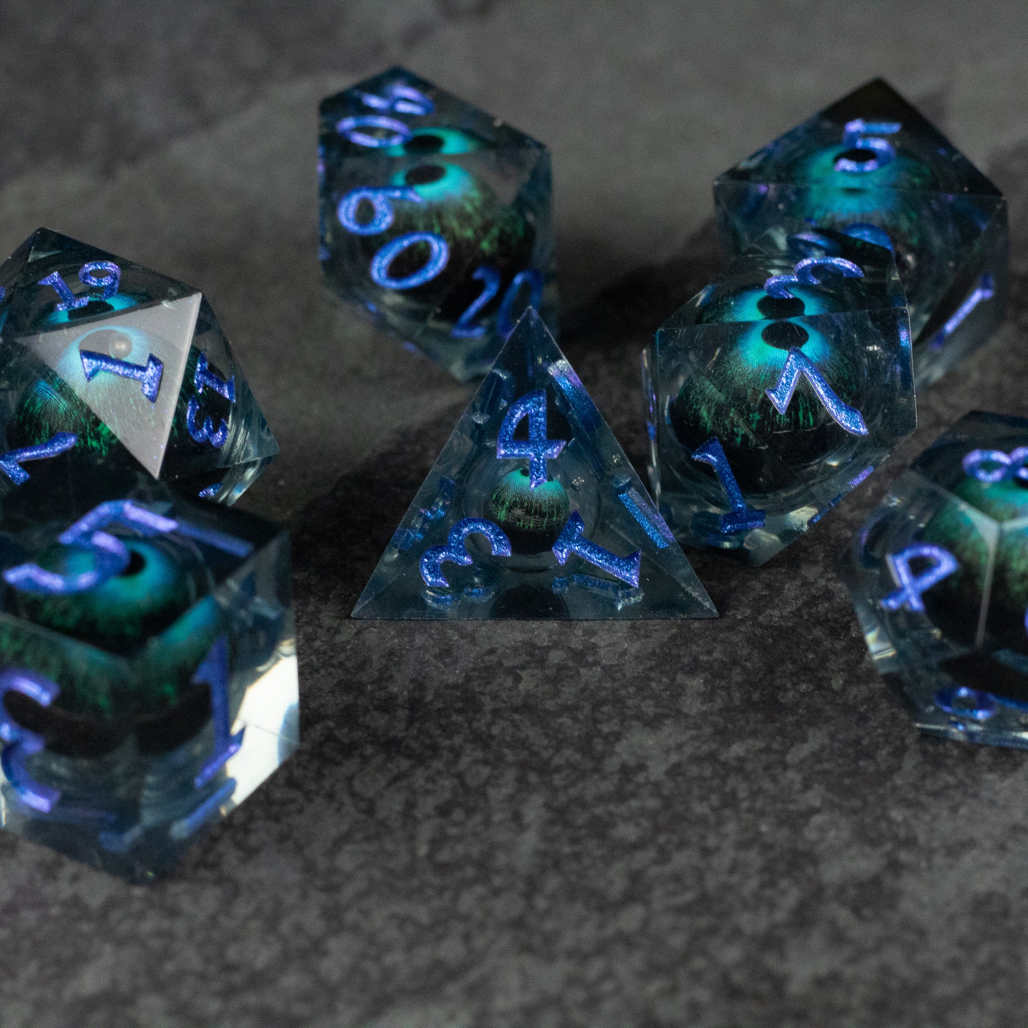 Blue Beholder's Eye Dice Set - Seven translucent blue and green polyhedral dice with sharp edges and glowing white numbers on a dark surface.