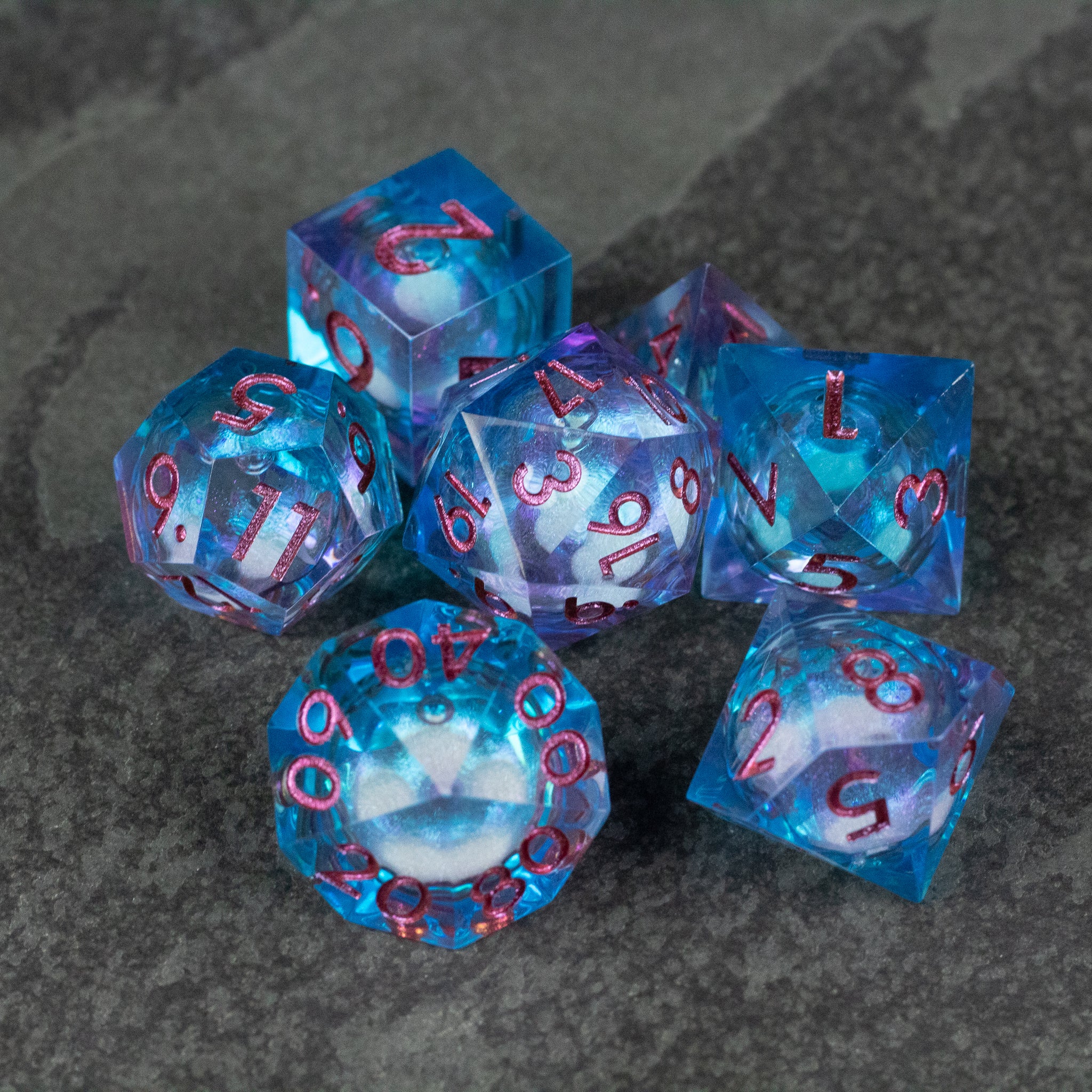 Crystallomancy Dice Set - Seven blue and purple liquid core polyhedral dice with sharp edges and red numbers on a dark stone surface.