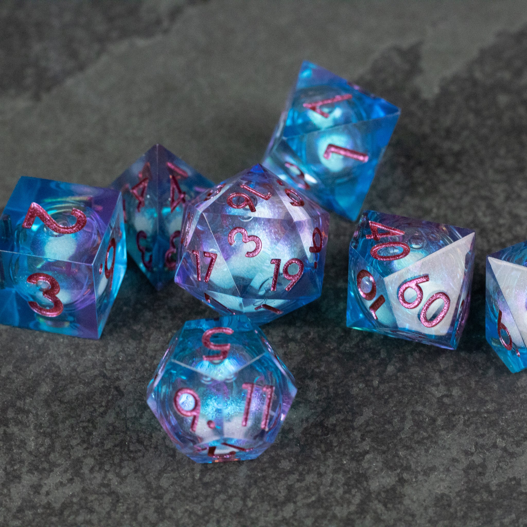 Crystallomancy Dice Set - Blue translucent polyhedral dice set with red numbers and liquid core effect on dark surface.