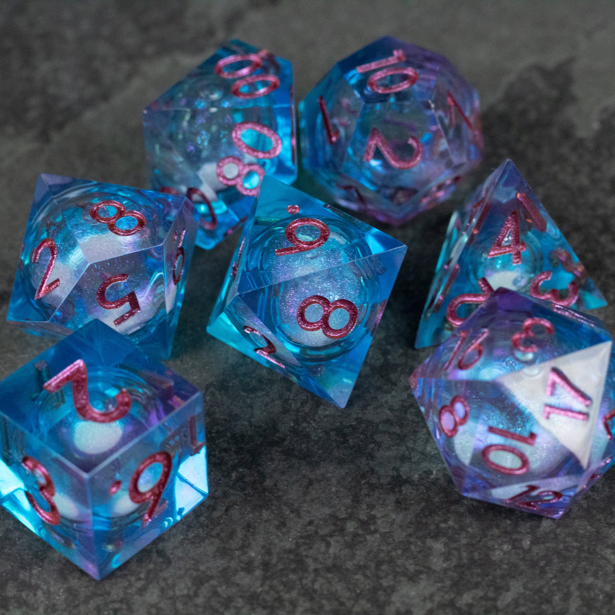 Crystallomancy Dice Set - Seven translucent blue polyhedral dice with pink numbers and a liquid core on a dark textured surface.