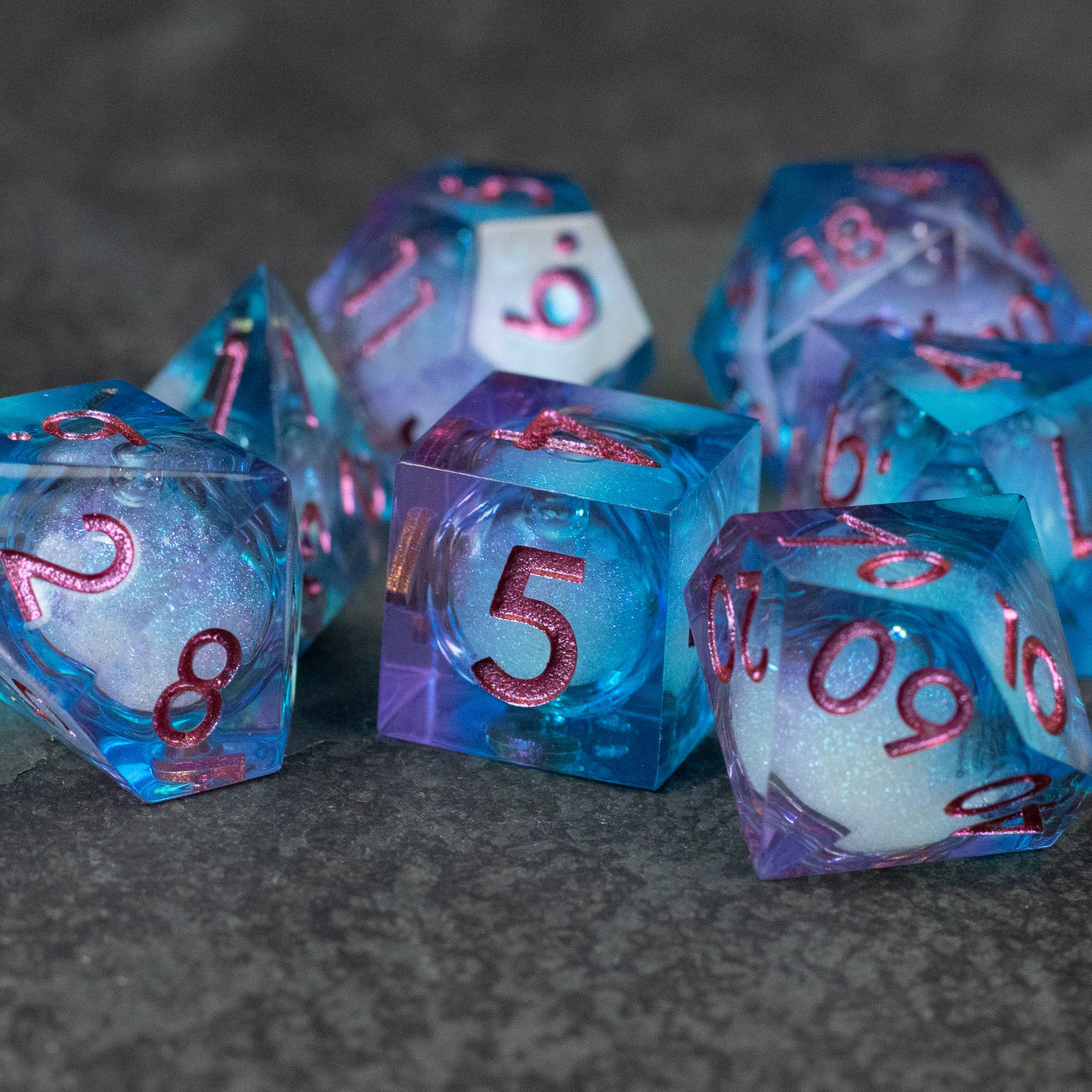Crystallomancy Dice Set - Set of translucent polyhedral dice with swirling blue, purple, and white liquid core and sharp-edged design, red numbers.
