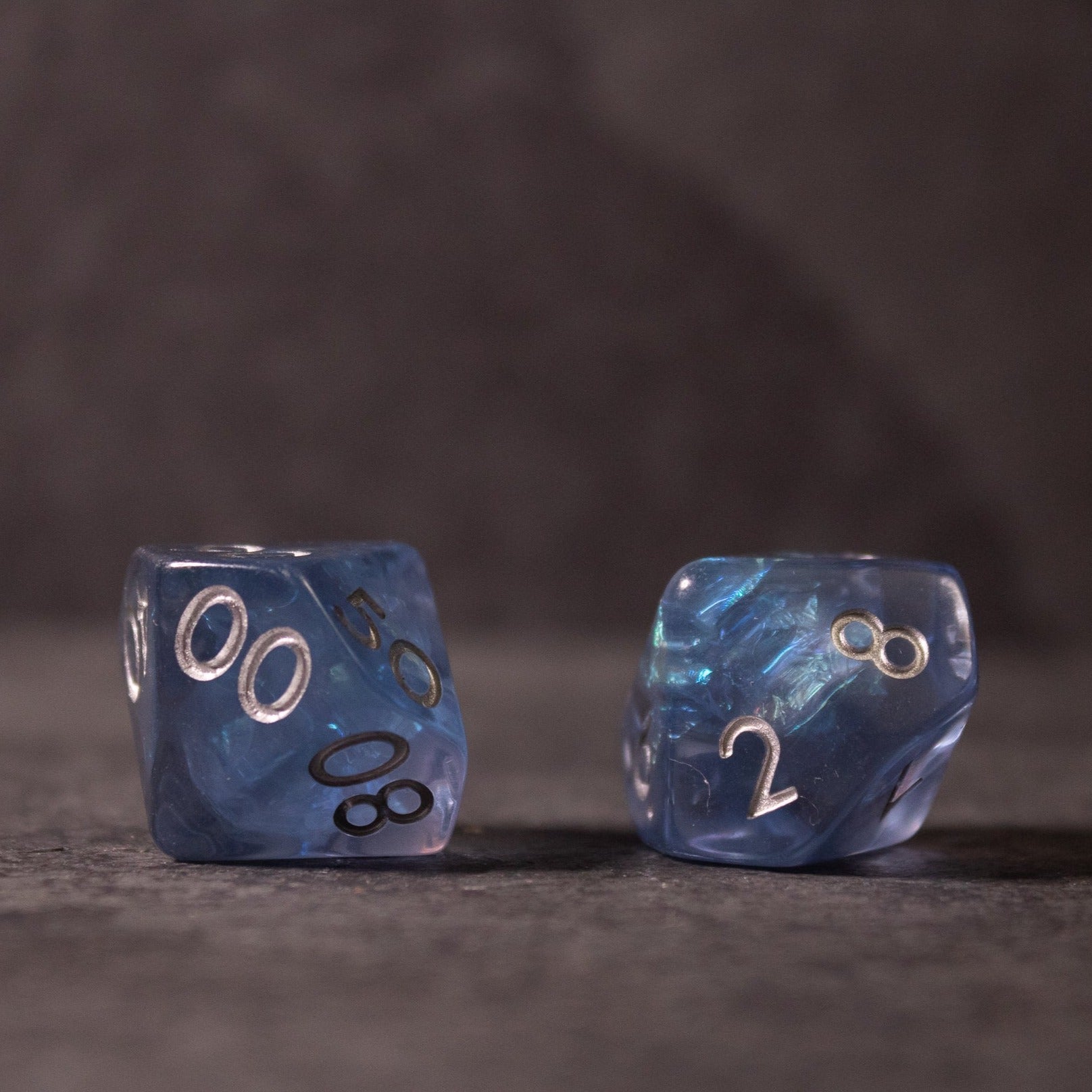 Glacier Ice Dice Set - Two blue iridescent d20 dice with silver numbering and swirl inclusions on a dark surface.