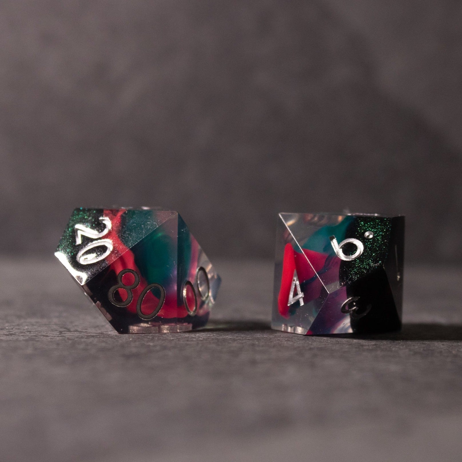 Pink & Green Flame Sharp Edge Dice Set - Two sharp-edged polyhedral dice with translucent pink and green swirls and white numbers on a dark surface.