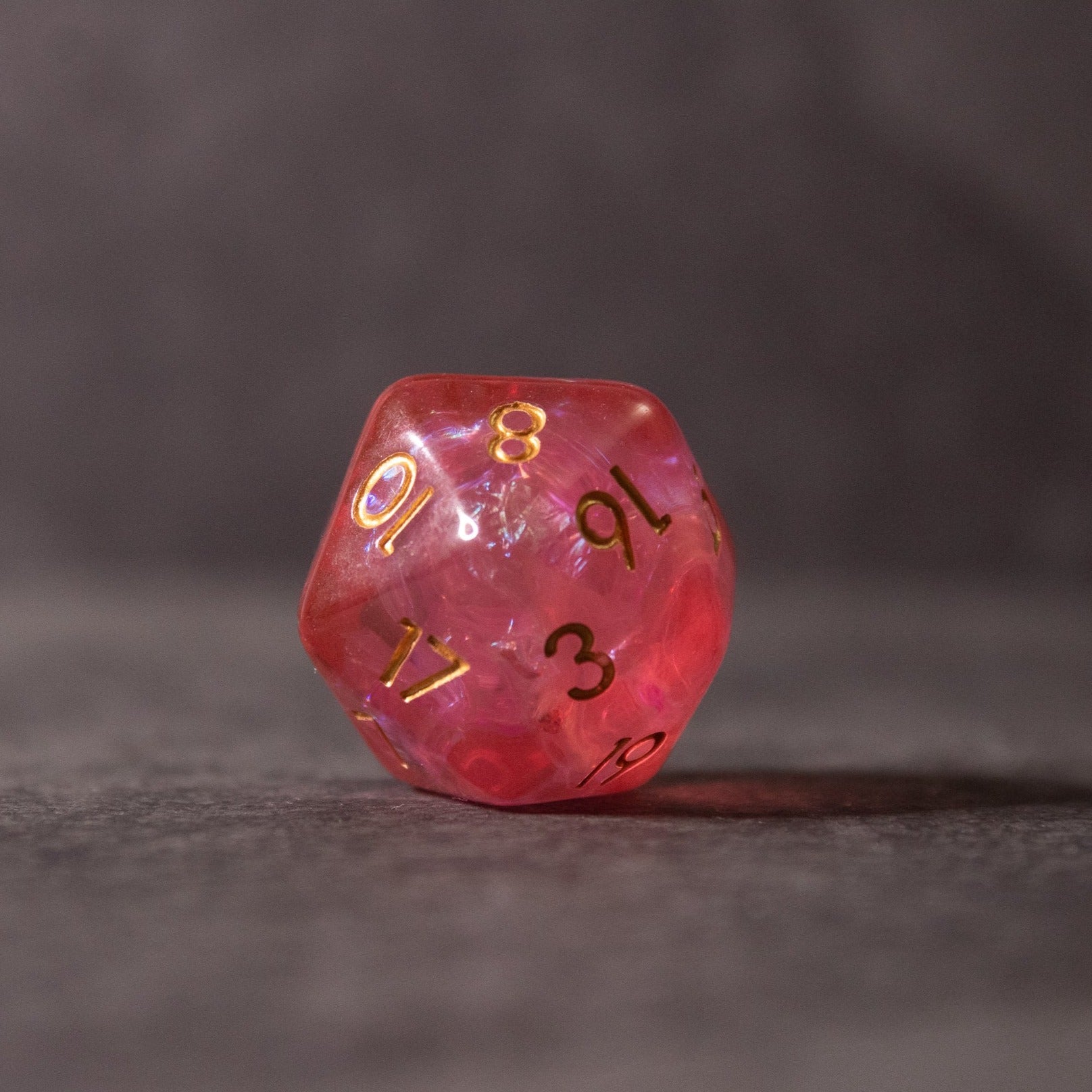 Pink Iridescent Dice Set - Pink iridescent 20-sided die with gold and black numbered faces on a dark surface.