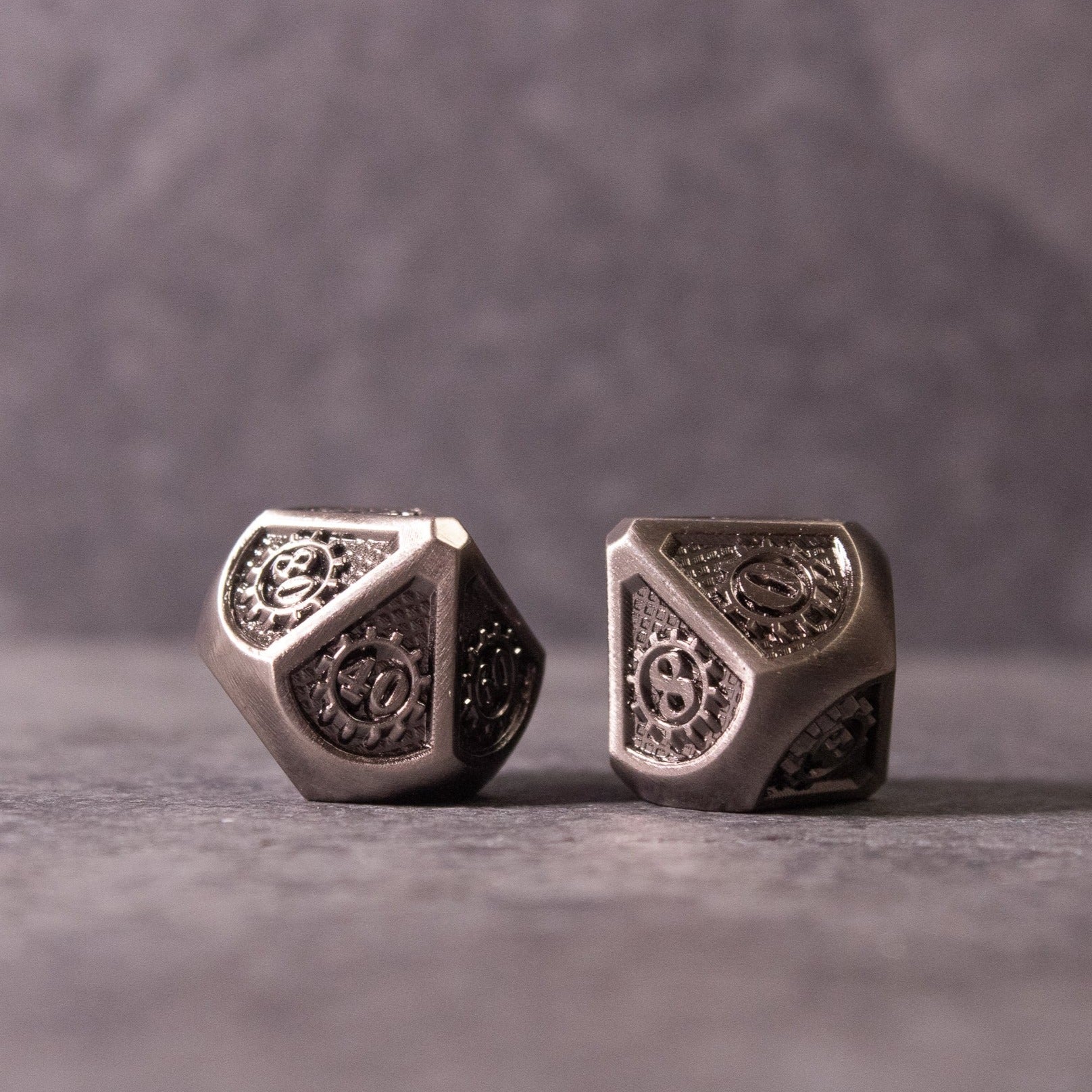 Metal Gear Dice Set - Two silver metal dice with intricate gear and cog designs on each face, reflecting light on a gray surface.