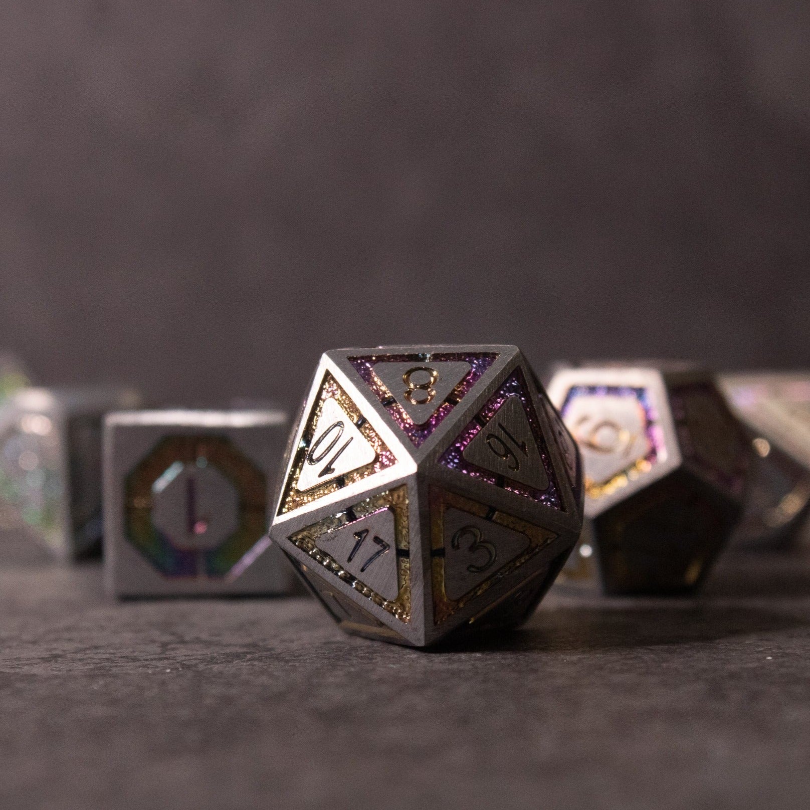 Prismatic Core Dice Set - Metallic d20 dice with prismatic core and etched numbers, other polyhedral dice blurred in background.