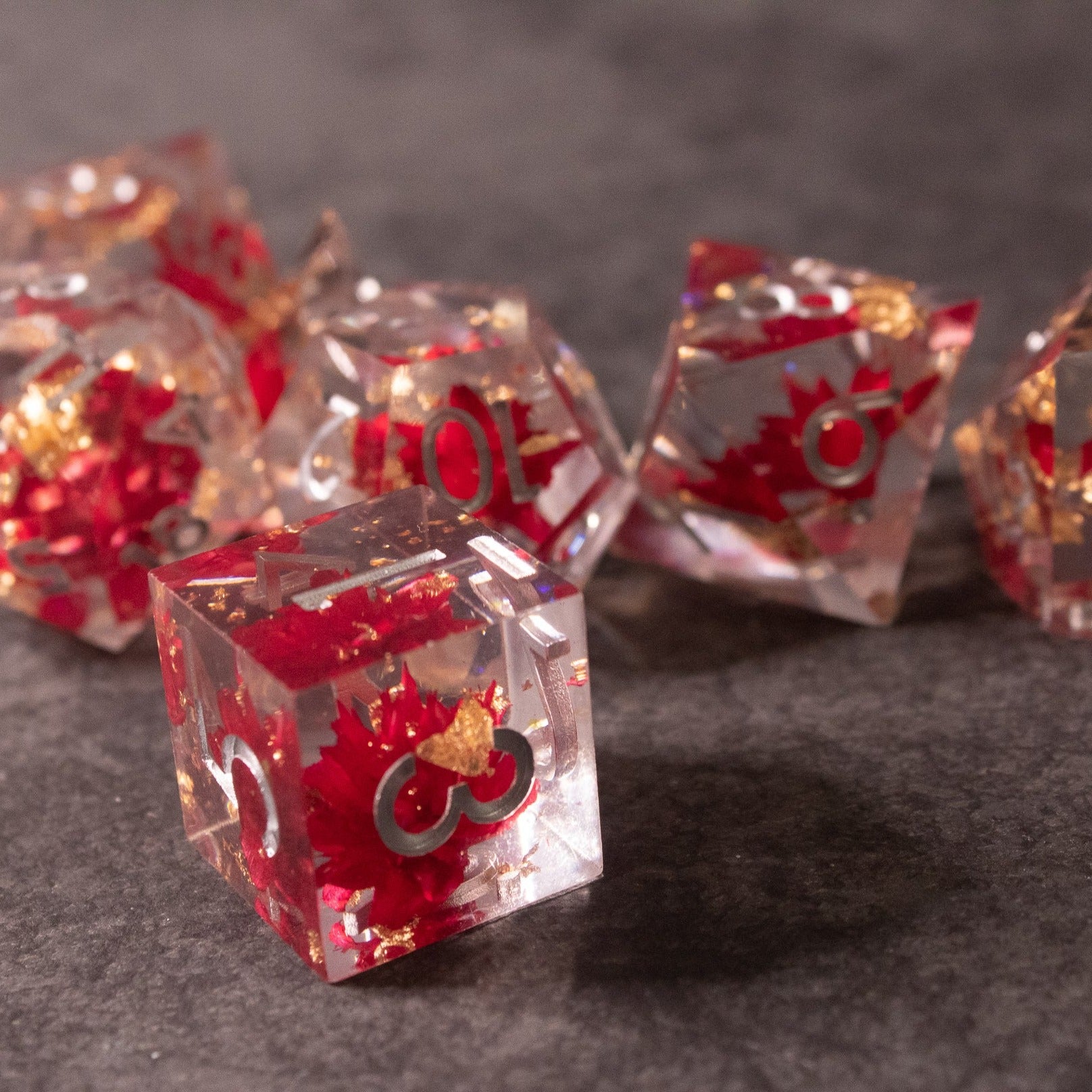 Red Lotus Flower Dice Set - Transparent sharp-edged dice with red lotus flower inclusions and red numbering on gray surface.
