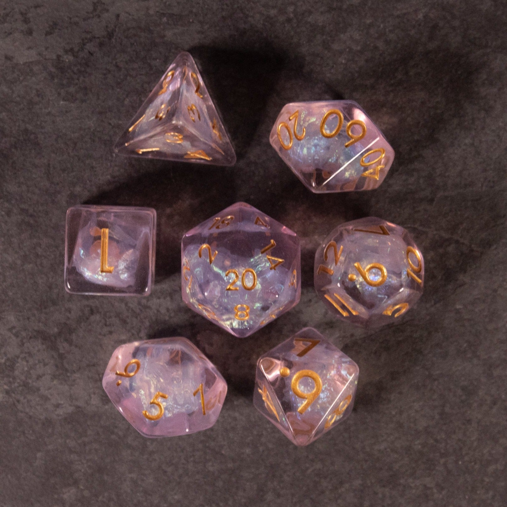 Sakura Dice Set - Seven translucent pink iridescent polyhedral dice with gold numbering on a dark surface.