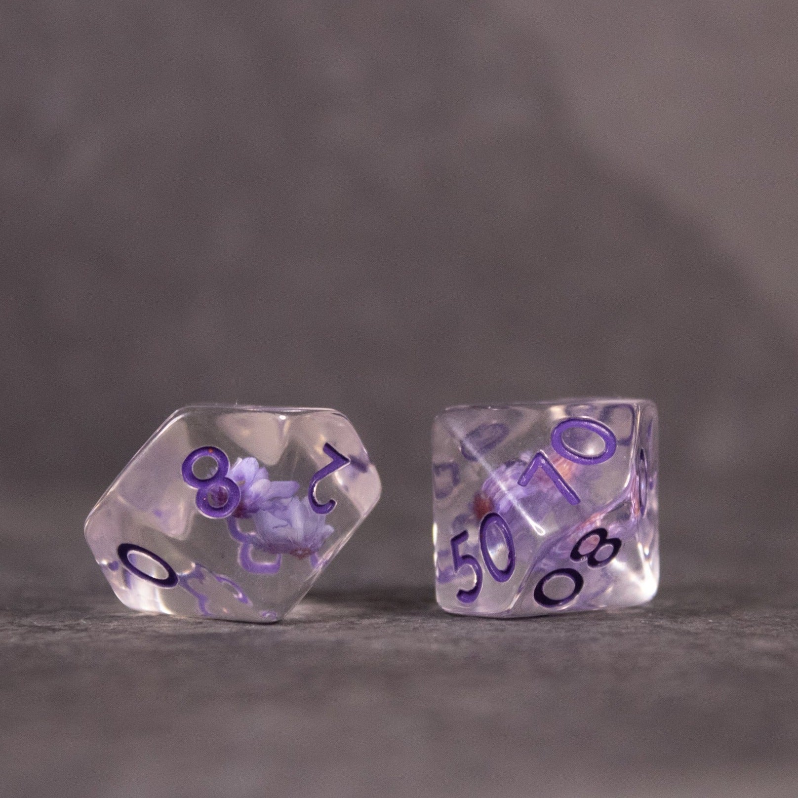 Purple Flower Dice Set - Two transparent purple dice with embedded purple flower inclusions and black numbering, one 10-sided and one 20-sided.