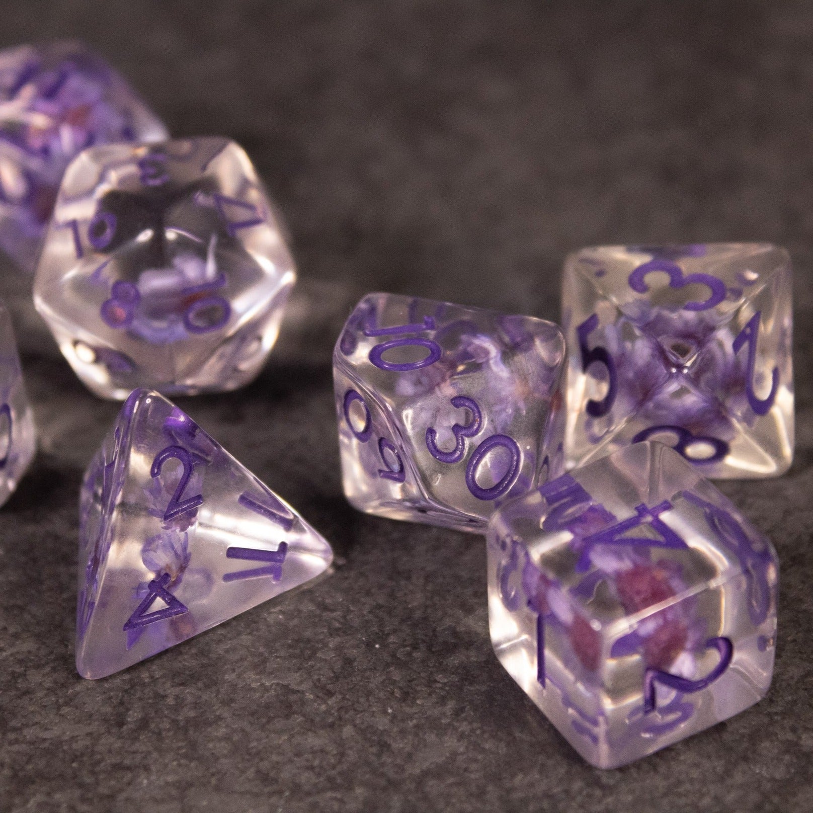 Purple Flower Dice Set - Transparent polyhedral dice set with purple numbers and embedded purple flower inclusions on a dark surface.